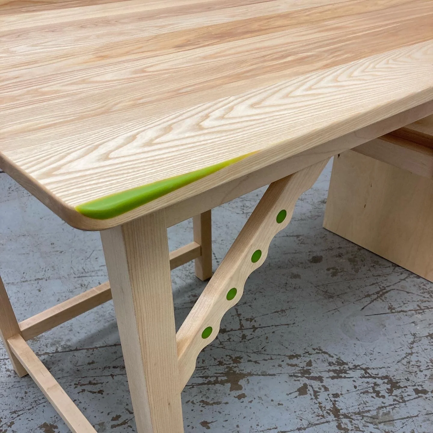Wooden table with a light-colored tabletop and a decorative green dot on the leg, set on a concrete floor.