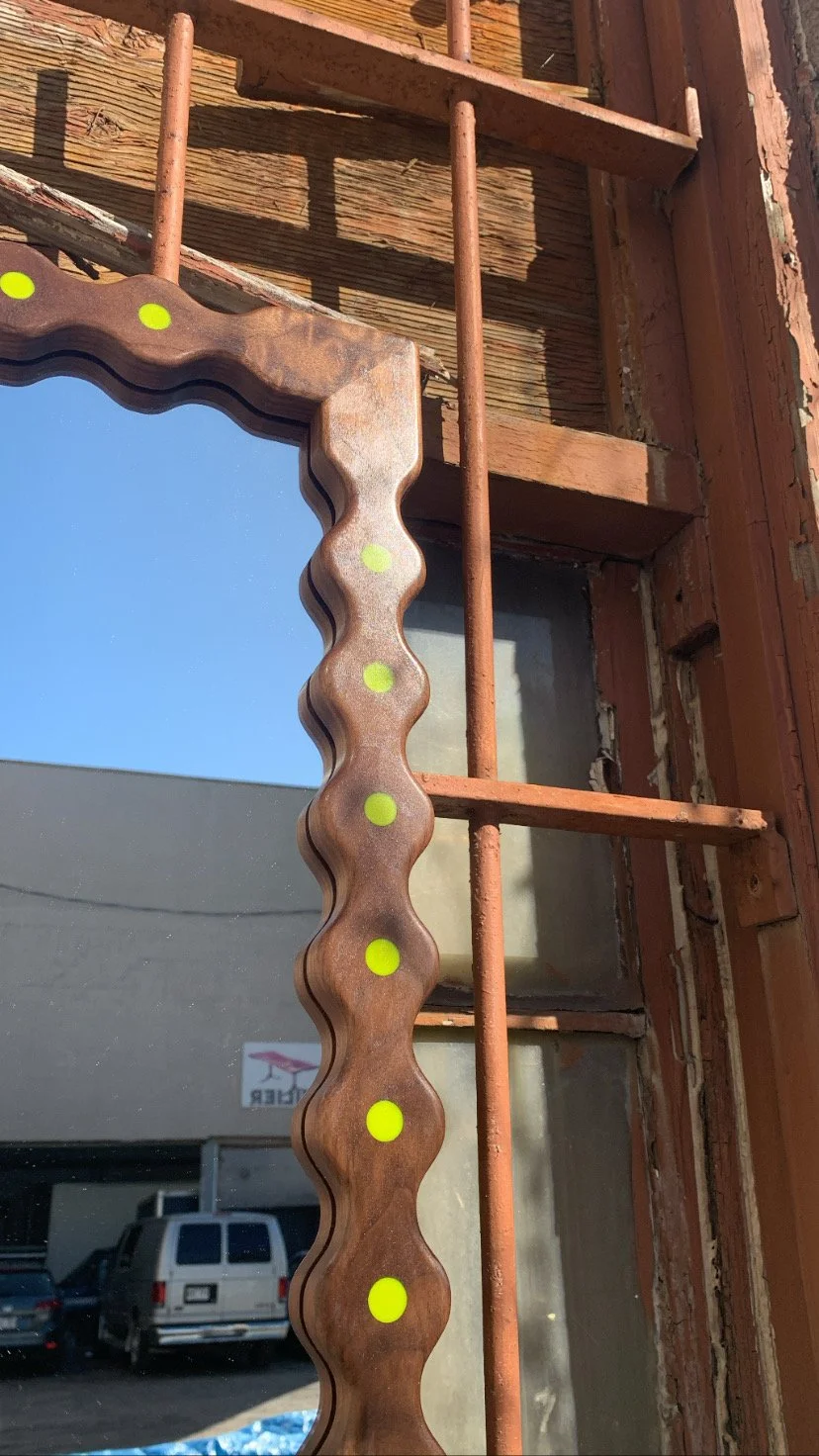 Close-up of a decorative wooden mirror frame with yellow dots, mounted on a window frame with rusty metal bars. The window area has weathered wood and peeling paint.