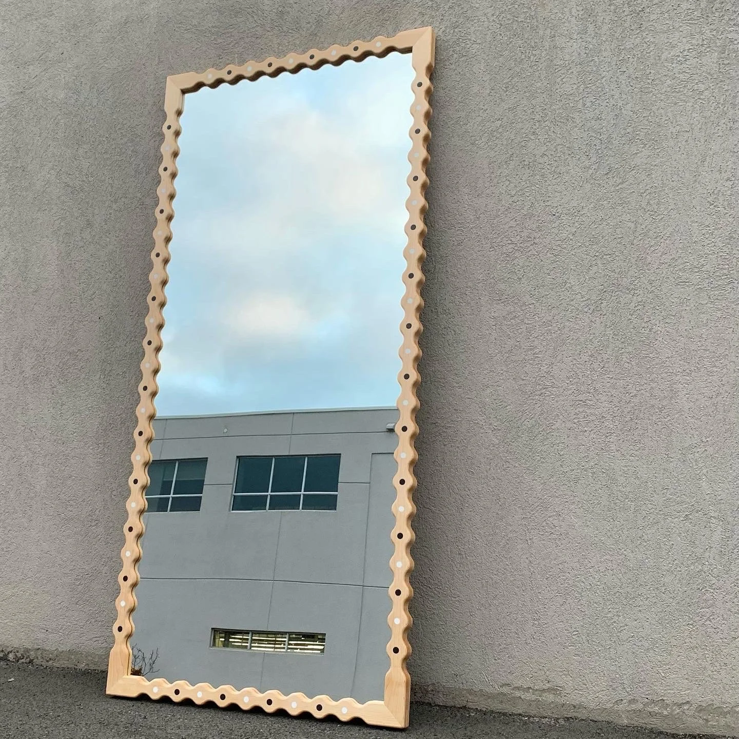 A tall, rectangular mirror with a wavy wooden frame leaning against a gray stucco wall. The mirror reflects a cloudy sky and part of a modern building with multiple windows.