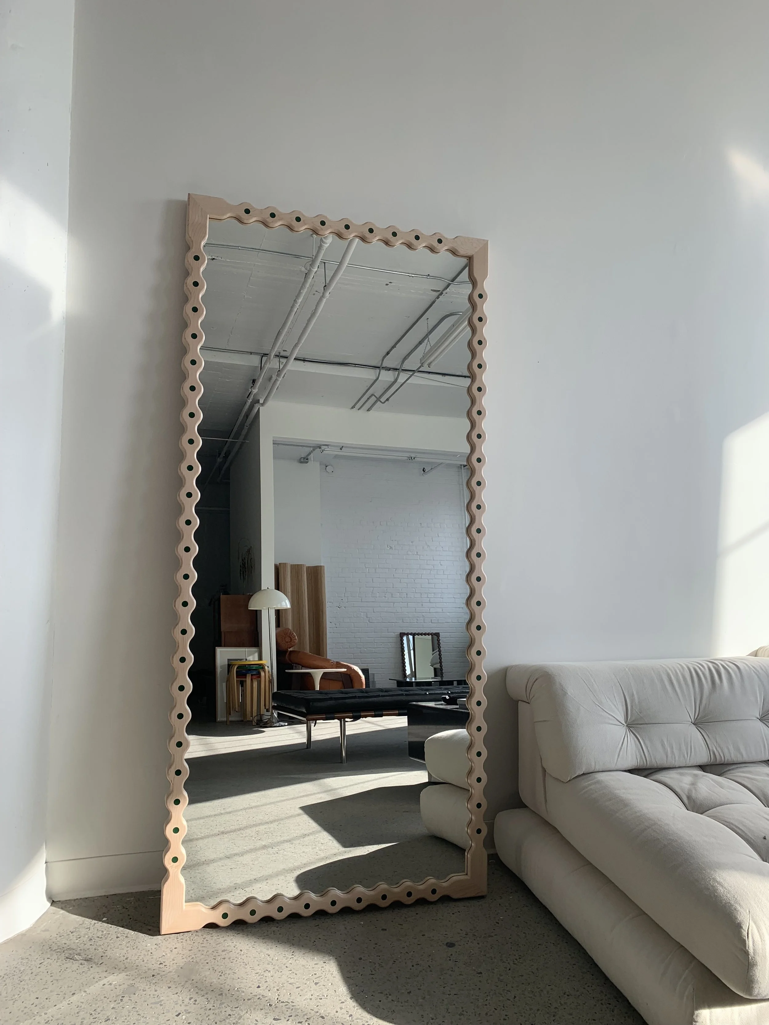 A large mirror with a wavy border leans against a white wall in a minimalist living room. The mirror reflects a white brick wall, a black bench, a white floor lamp, a small framed mirror, and some wooden furniture in the background. A cream-colored s