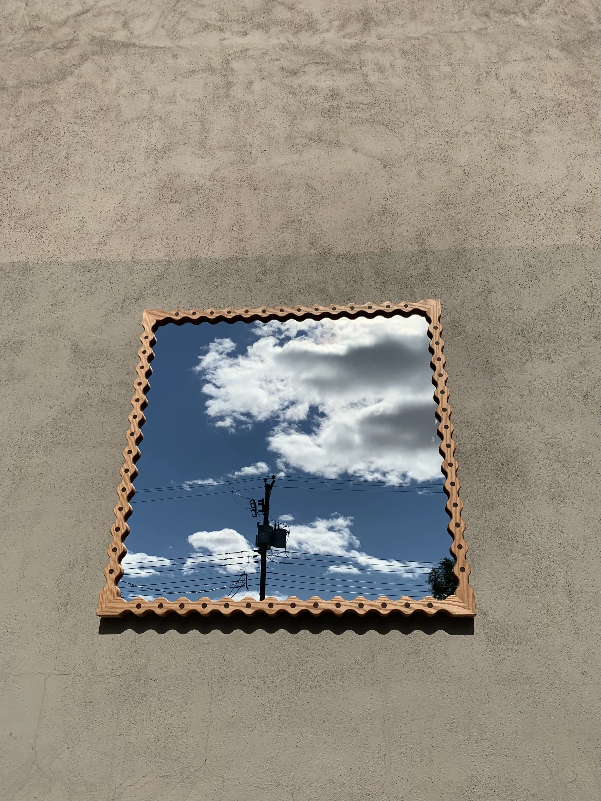 A square mirror with a zigzag wooden frame reflecting a blue sky with scattered white clouds and an electric pole with wires.
