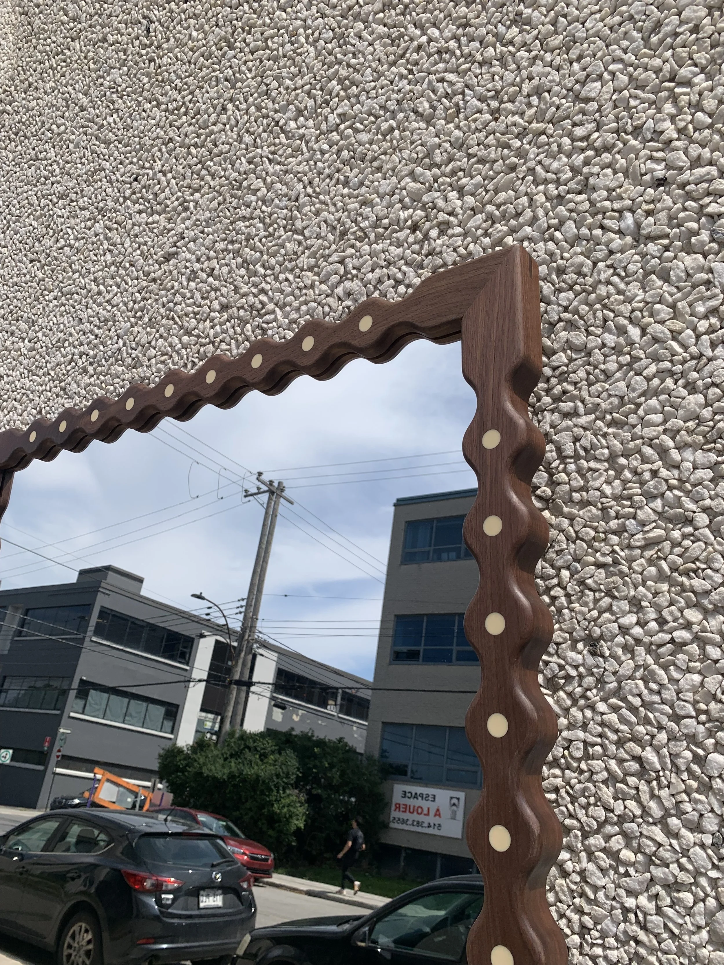 A mirror with a wavy wooden frame mounted on a textured wall. The mirror reflects city buildings, a street lamp, power lines, a person walking, and parked cars.