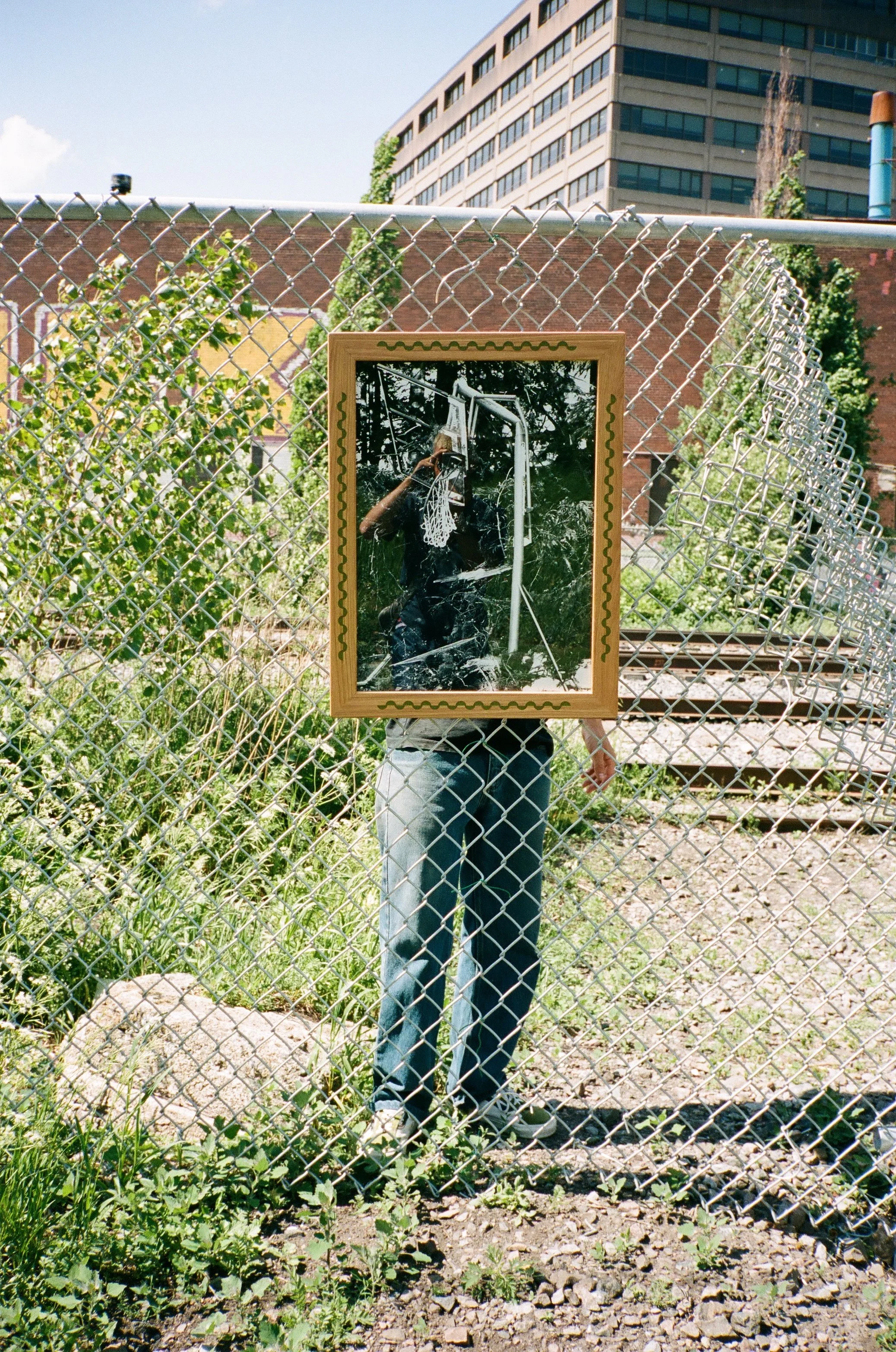 A person standing behind a chain-link fence, holding a framed black-and-white photo of a person dunking a basketball. The real person is reflected in the glass of the framed photo, creating the illusion that they are the person dunking.