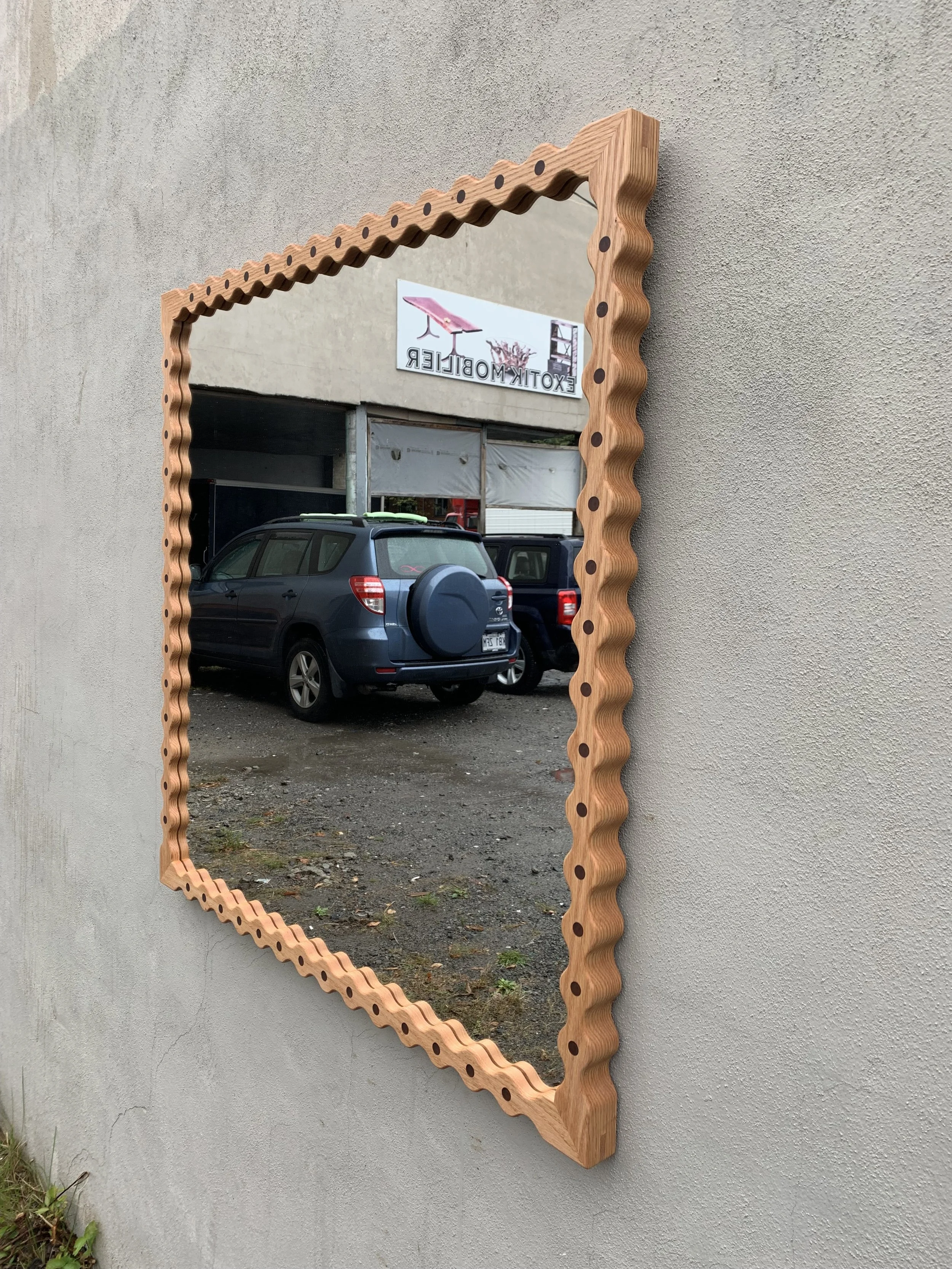 Decorative wall mirror with a wavy wooden frame mounted on a light-colored exterior wall, reflecting a parking area with vehicles and a sign that reads 'EOTIK MOBILER'.