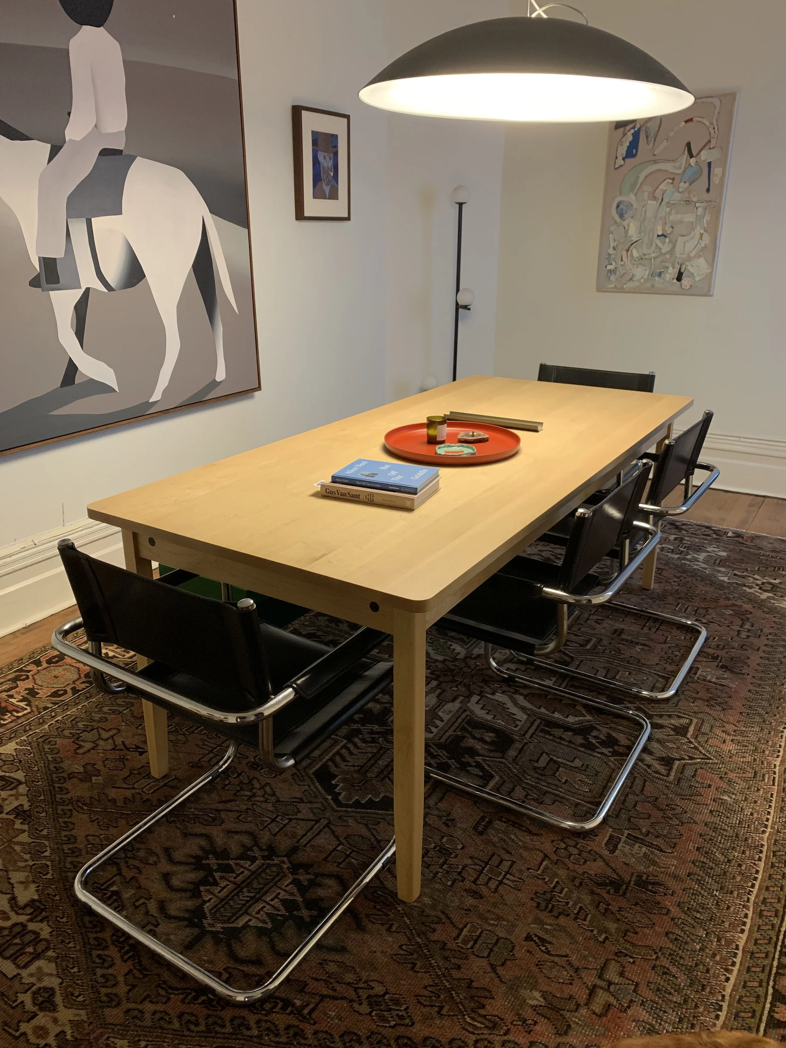 Room with a wooden dining table, four black chairs, a colorful painting on the wall, a large overhead light, a patterned rug, and some books and items on the table.