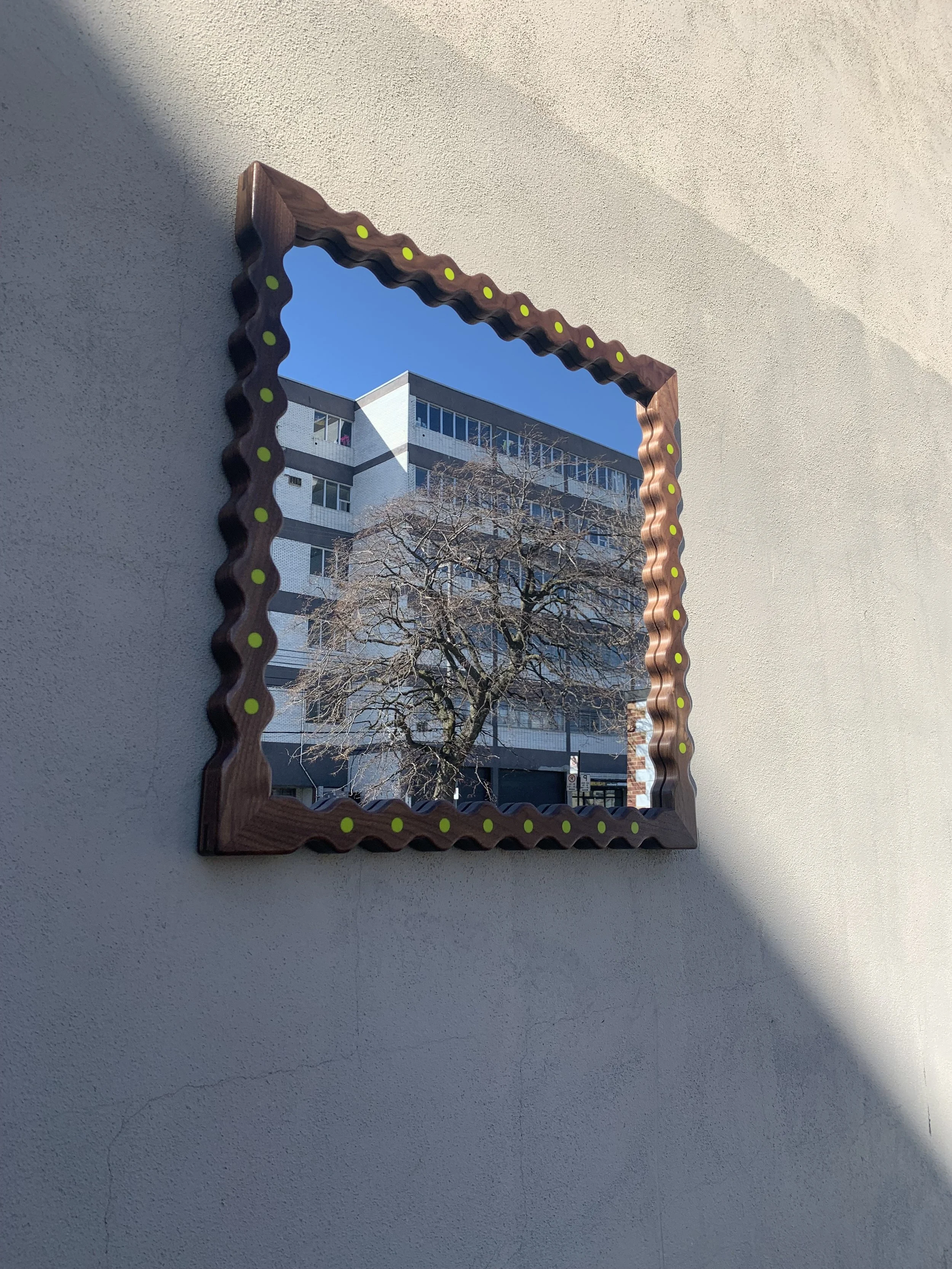 A mirror with a decorative wooden frame featuring small yellow dots, mounted on a beige wall. The mirror reflects an outdoor scene with a leafless tree and a modern multi-story building against a clear blue sky.