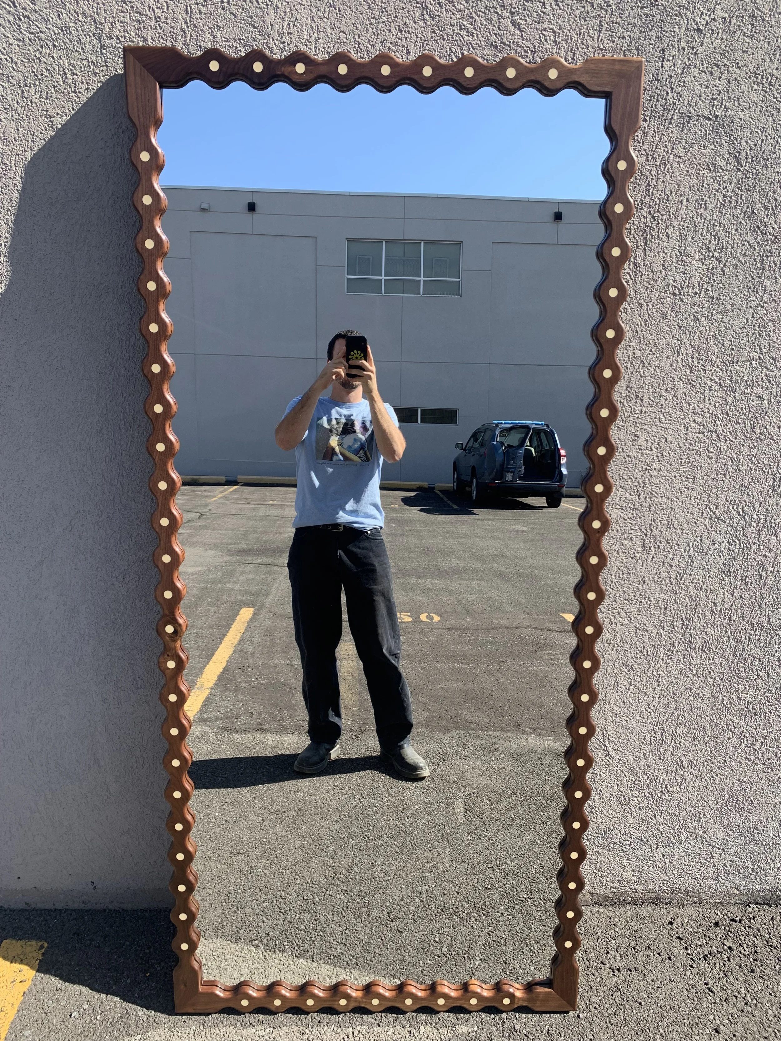 Person taking a selfie in a decorative mirror mounted on a wall in a parking lot on a sunny day.
