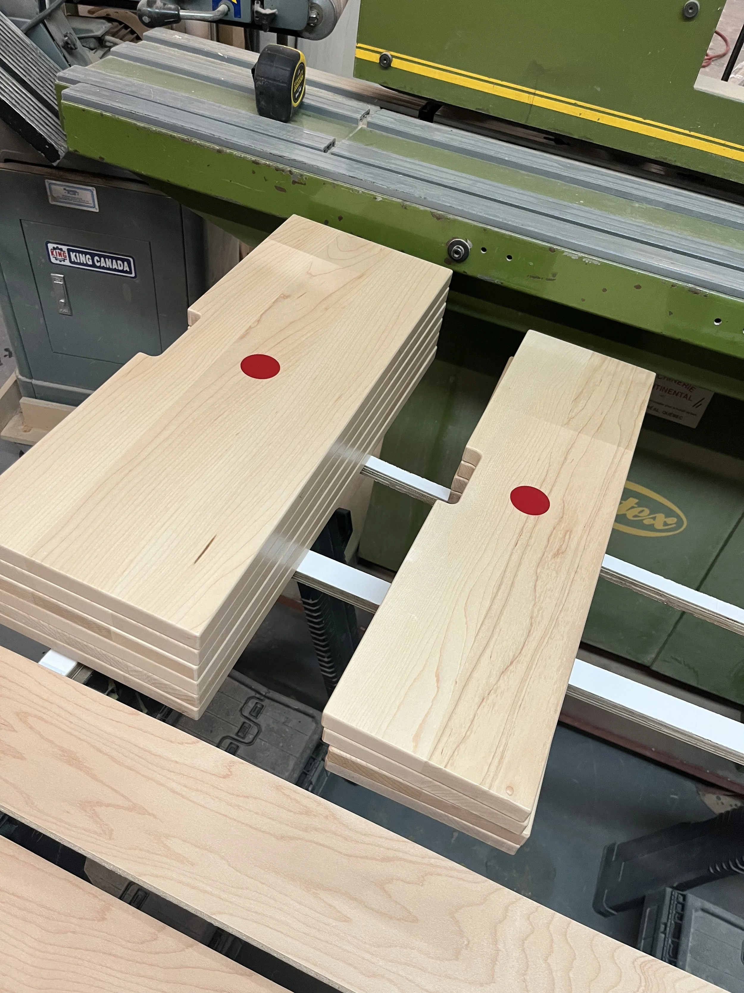 Several light-colored wooden planks with red circular stickers on top, placed on a workbench with a green woodworking machine in the background.