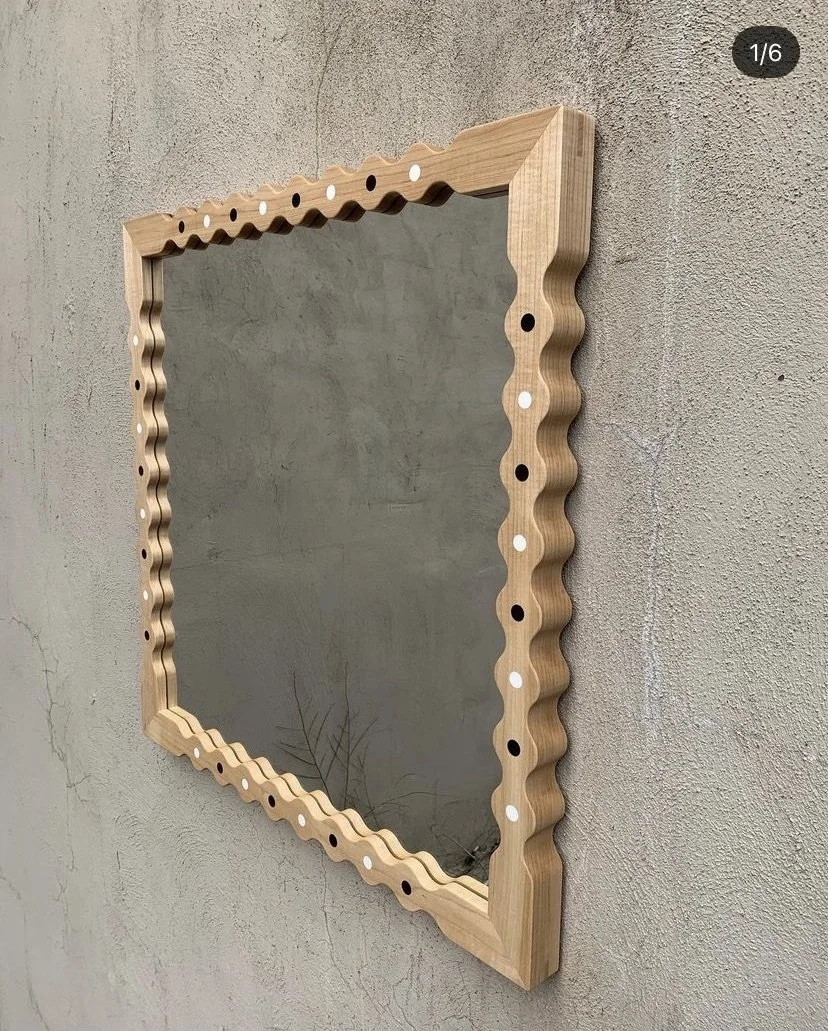 A rectangular wall mirror with a wavy wooden frame decorated with alternating black and white circular dots, mounted on a textured beige wall.