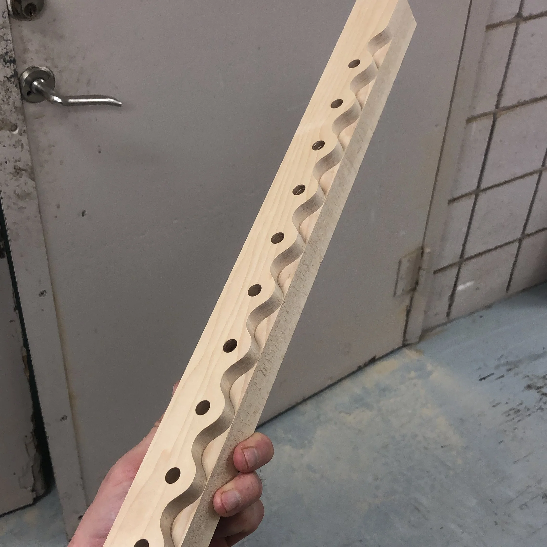A person holding a wooden piece with a wavy, grooved pattern and round holes along the side, in front of a gray door and a cinder block wall.