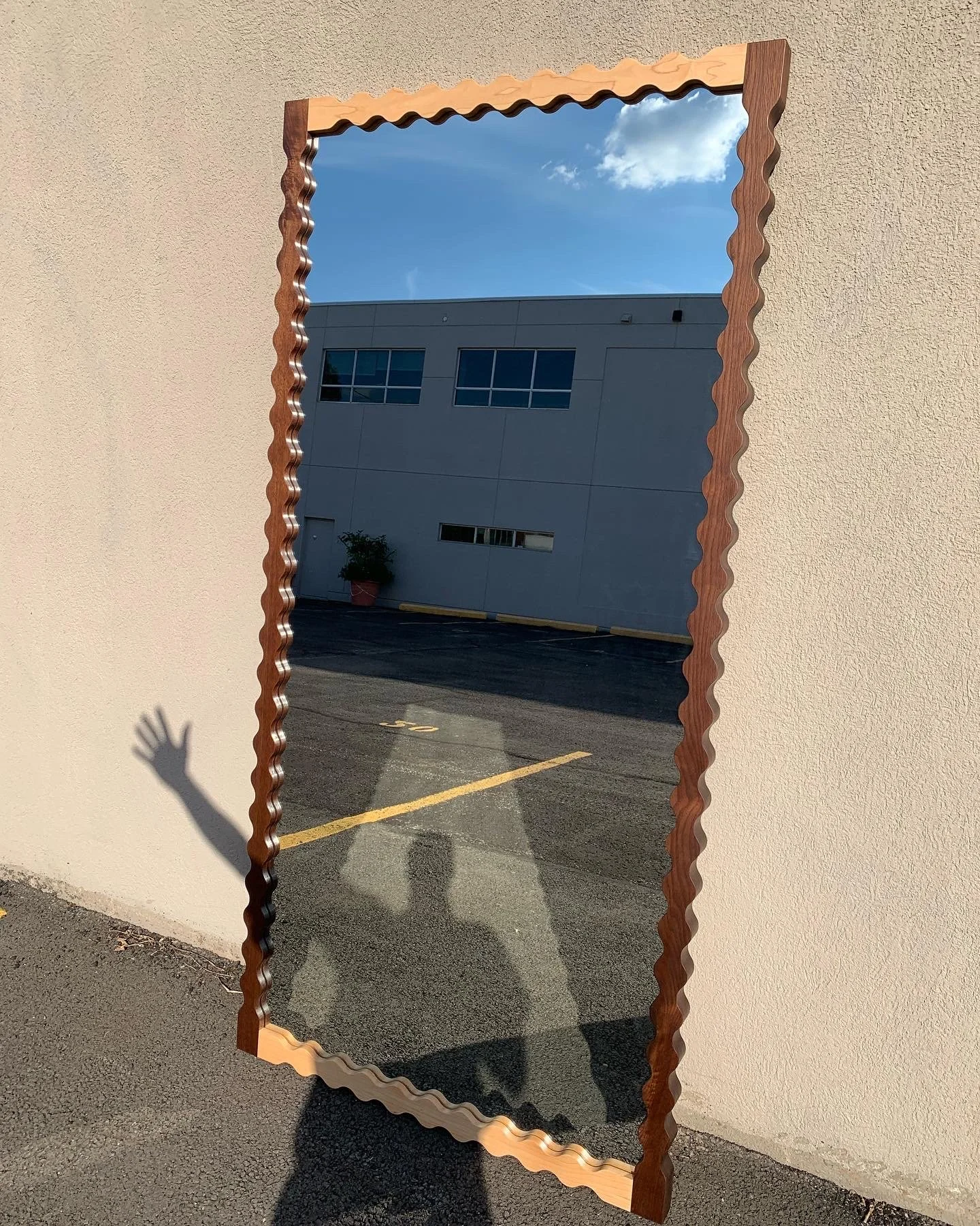 A rectangular mirror with a wavy wooden frame leaning against a beige wall, reflecting a building, blue sky with clouds, and a parking lot.