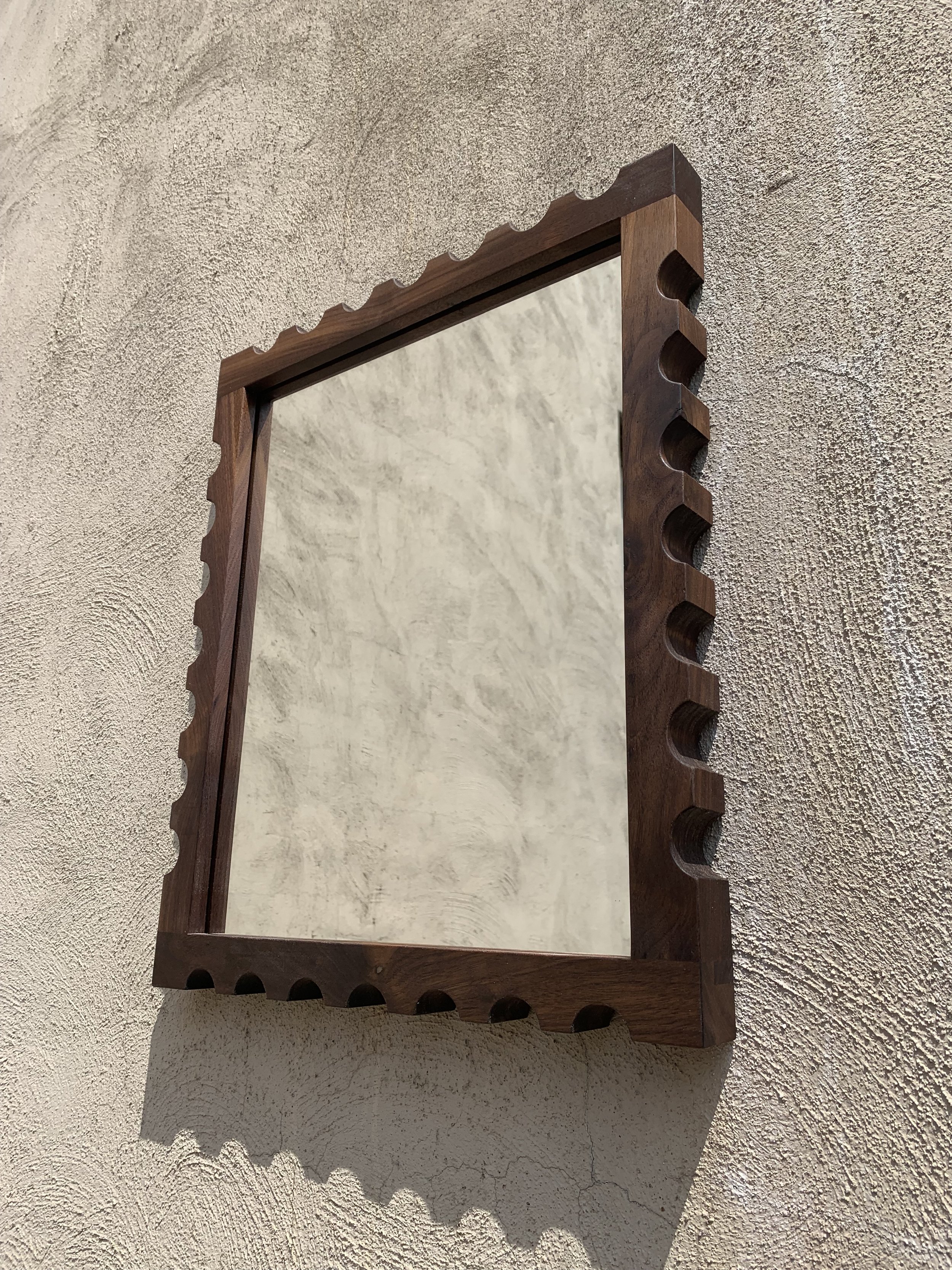A square mirror with a wooden frame that has a scalloped edge design, mounted on a textured beige wall.