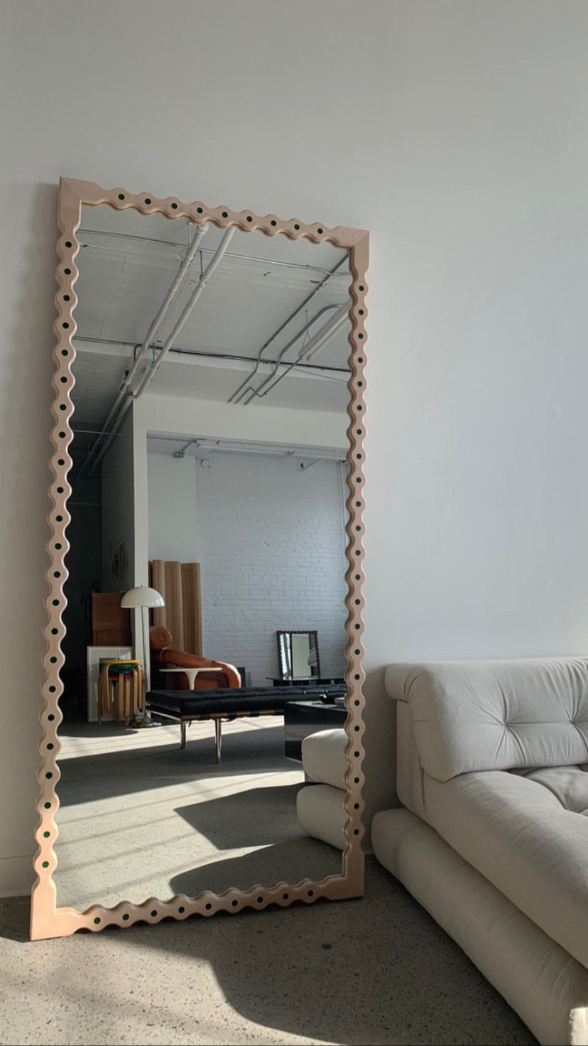 a tall mirror with a wavy beige frame, reflecting a modern interior space with white walls, a white brick wall in the background, industrial ceiling pipes, a beige chair, a black table, and a white ceiling lamp.