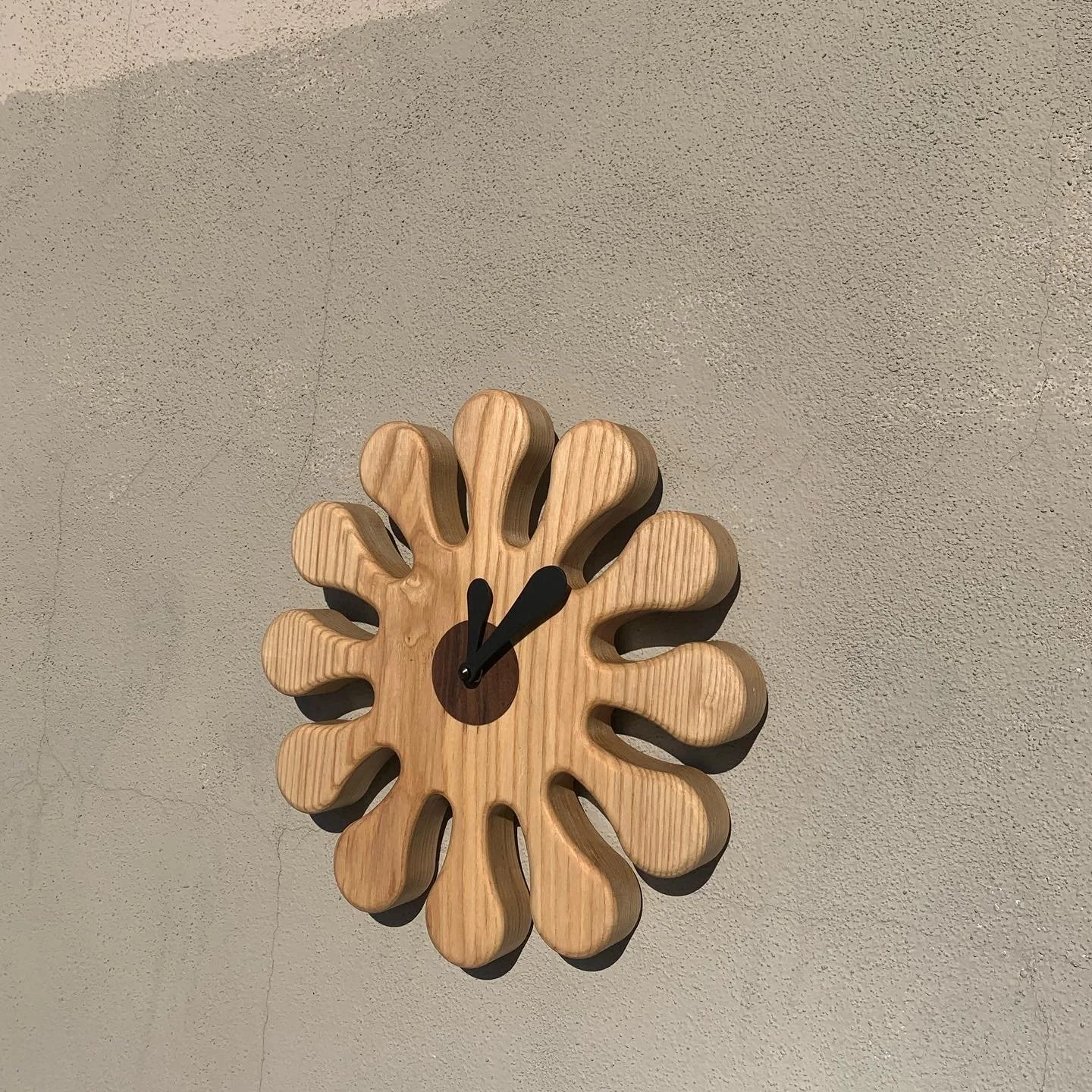 Wooden wall clock with petal-shaped design and black clock hands on a textured beige wall.