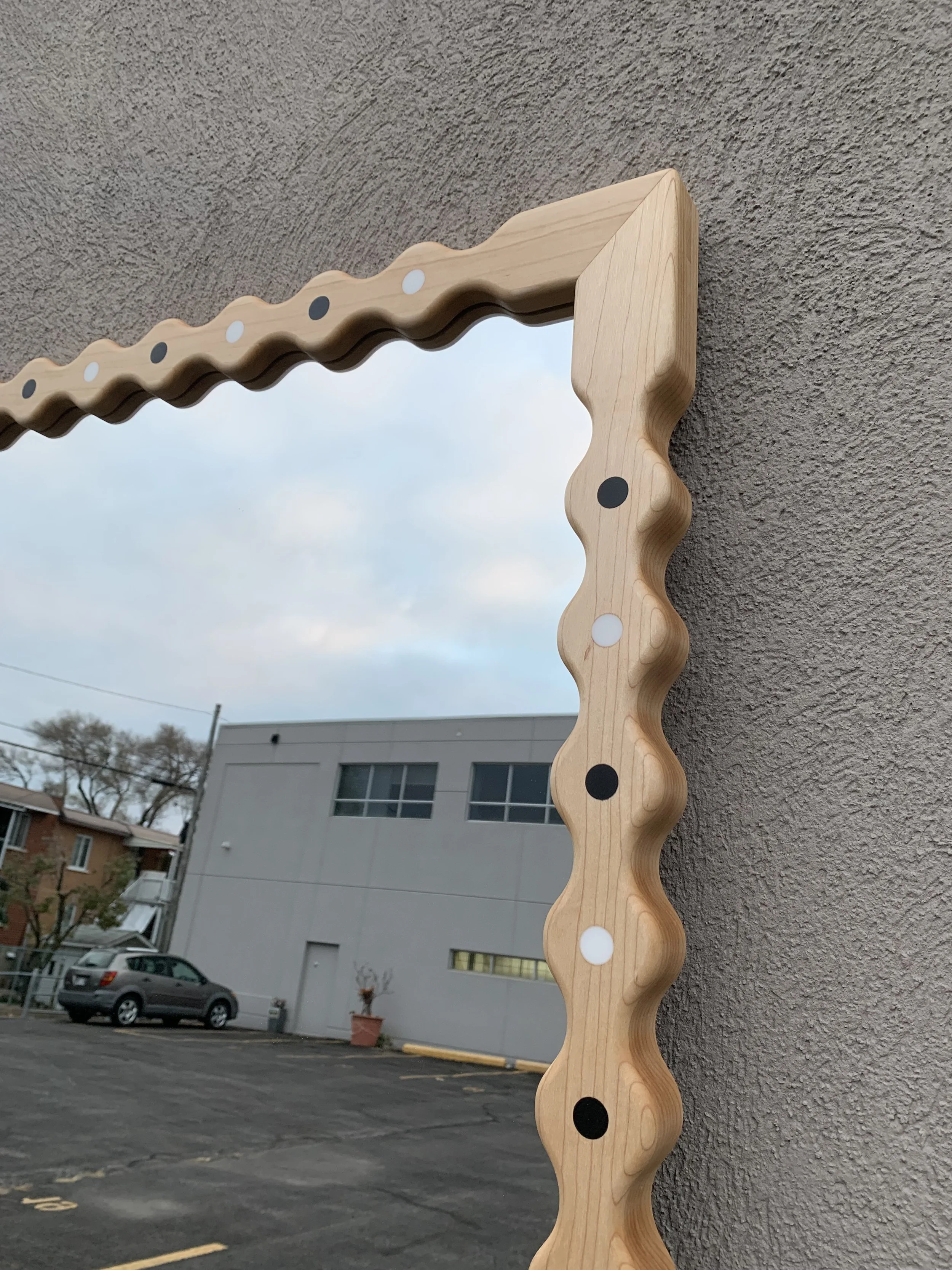 Decorative mirror with a wavy, light wood frame mounted on an outdoor wall, reflecting a cloudy sky.