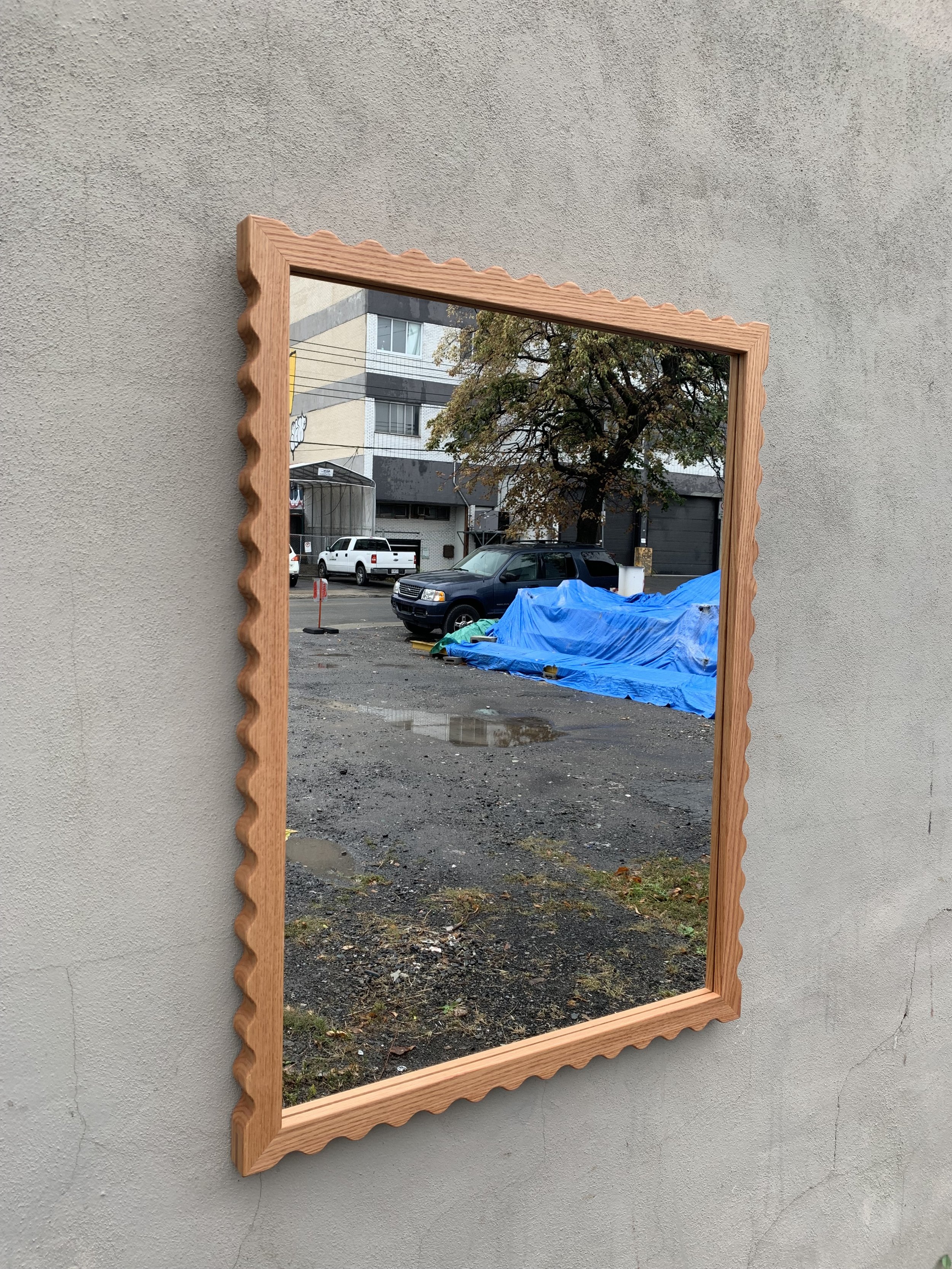 A rectangular mirror with a scalloped wooden frame hanging on a plain beige wall, reflecting an outdoor scene of parked cars, a tree, and a blue tarp covering an object on a gravel lot.