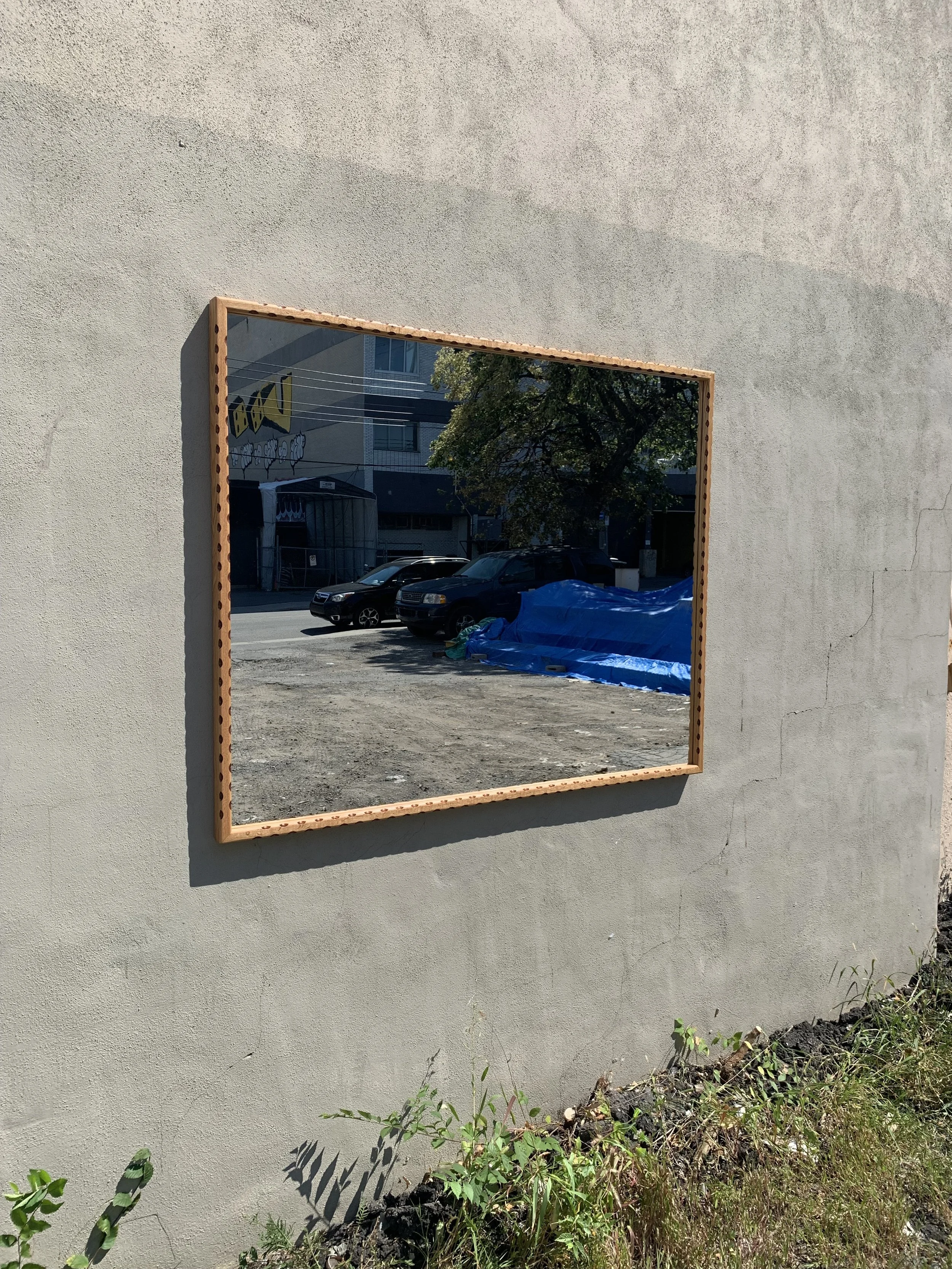 A rectangular mirror with a wooden frame mounted on a beige exterior wall reflecting a street scene with parked cars, a building, and a tree.