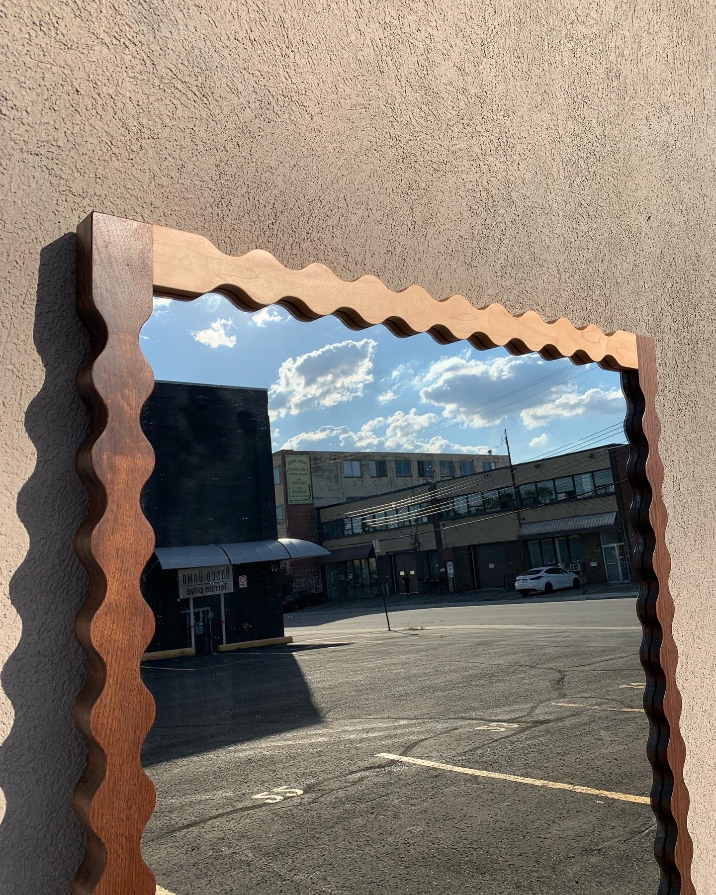 Reflection of a city street scene with buildings, parked cars, power lines, and a cloudy sky in a decorative wooden-framed mirror hanging on a beige textured wall.