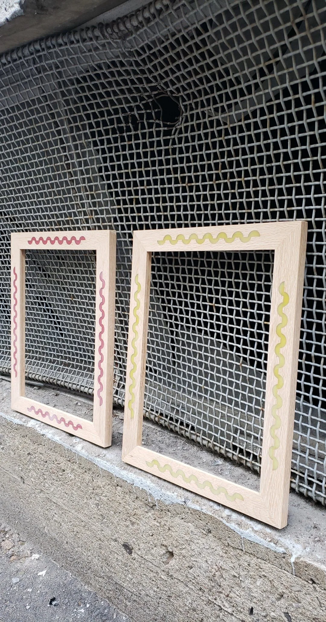Two wooden picture frames with wavy painted borders, one pink and one yellow, leaning against a wire mesh fence.