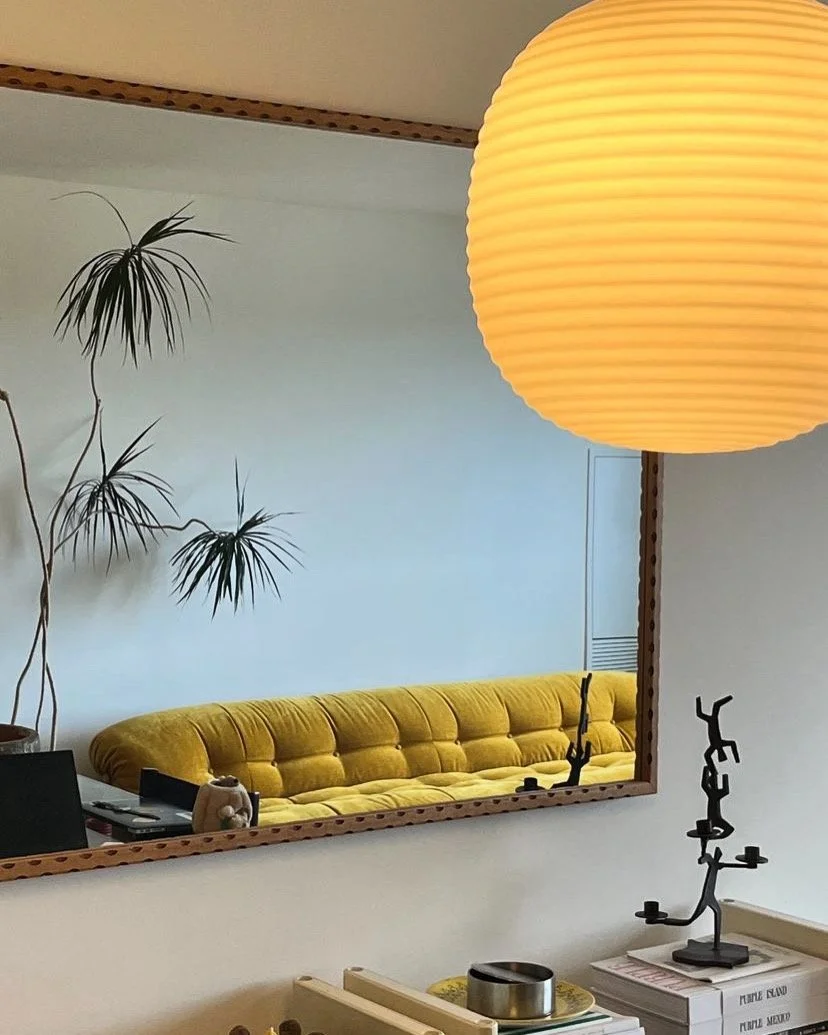 Living room with a large mirror reflecting a yellow tufted sofa, a potted plant, a laptop on a side table, and a paper lamp hanging from the ceiling.