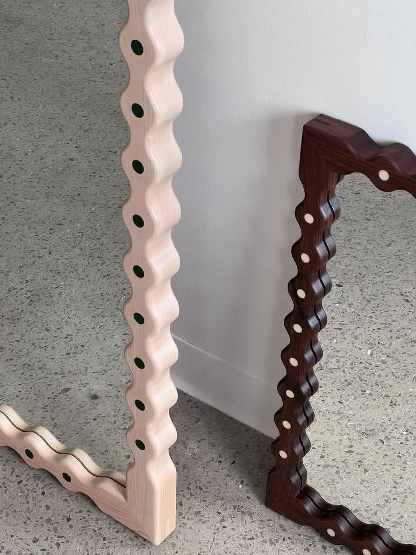 Close-up of two picture frames with wavy edges, one light wood and one dark wood, leaning against a white wall on a gray speckled floor.