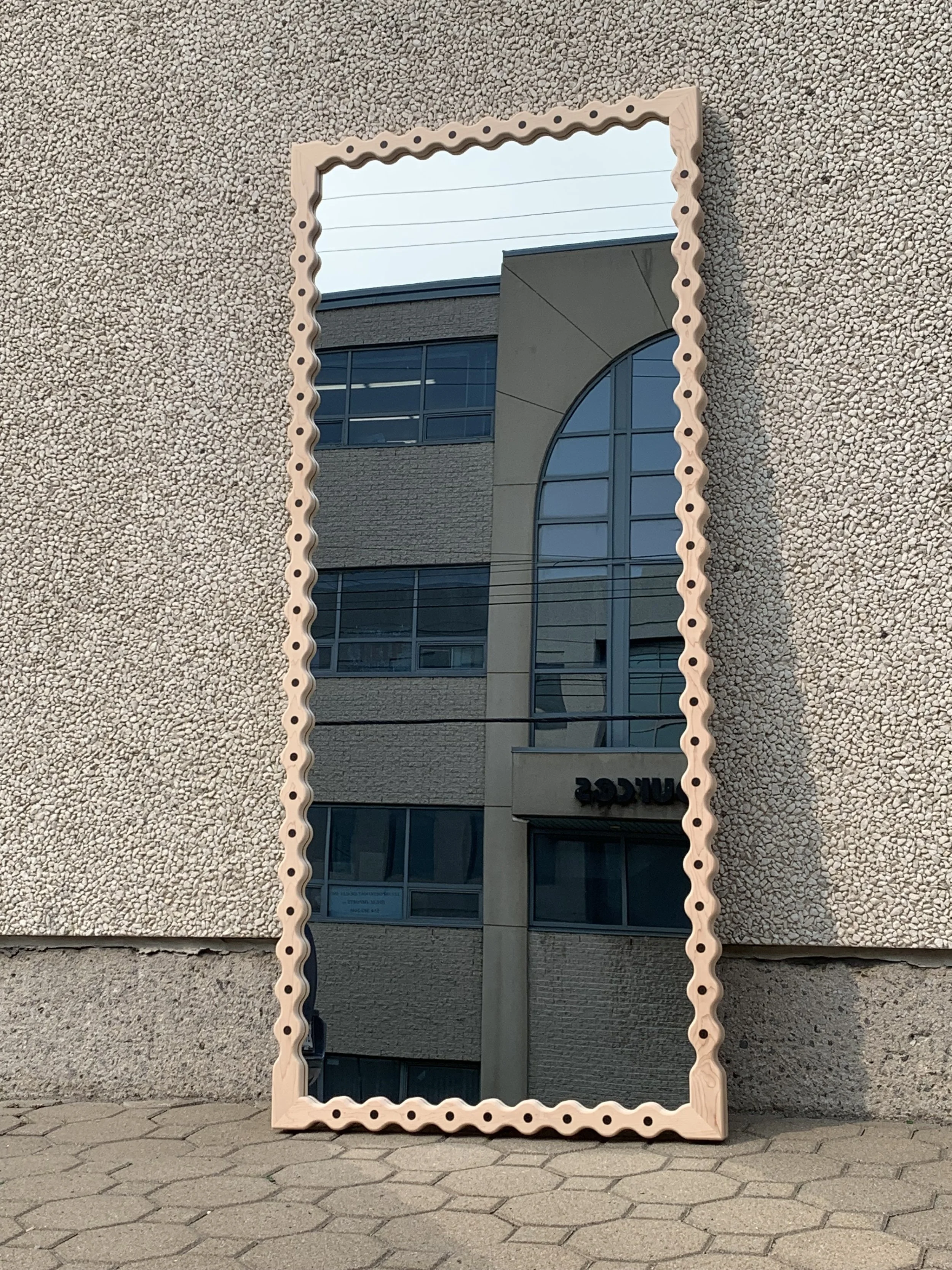 A tall rectangular mirror with a decorative wooden frame featuring rounded cutouts, leaning against a textured exterior wall of a building, reflecting a multi-story modern building with large glass windows and a curved architectural feature.