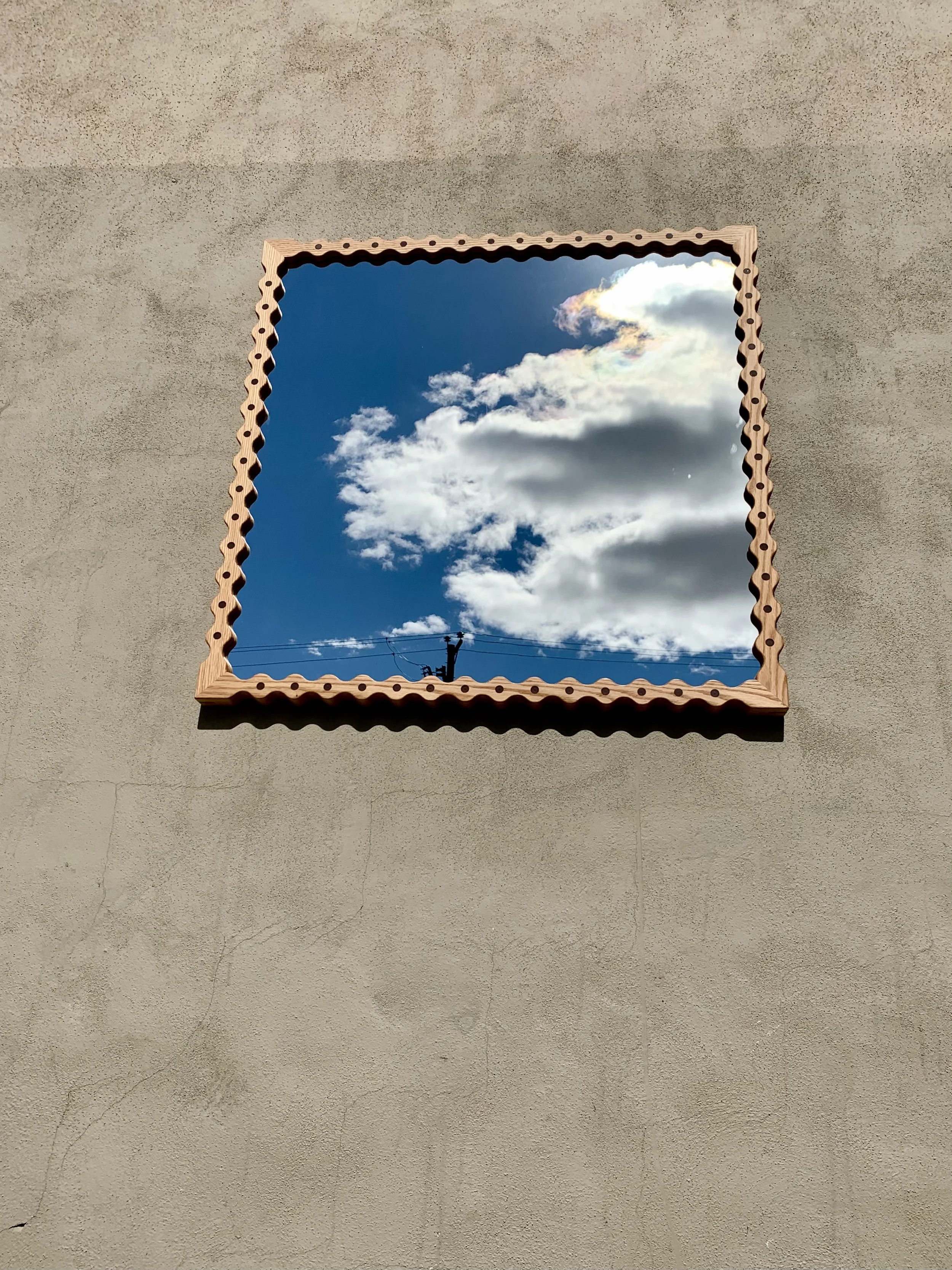 A mirror on a beige wall reflects a blue sky with clouds and a power line.