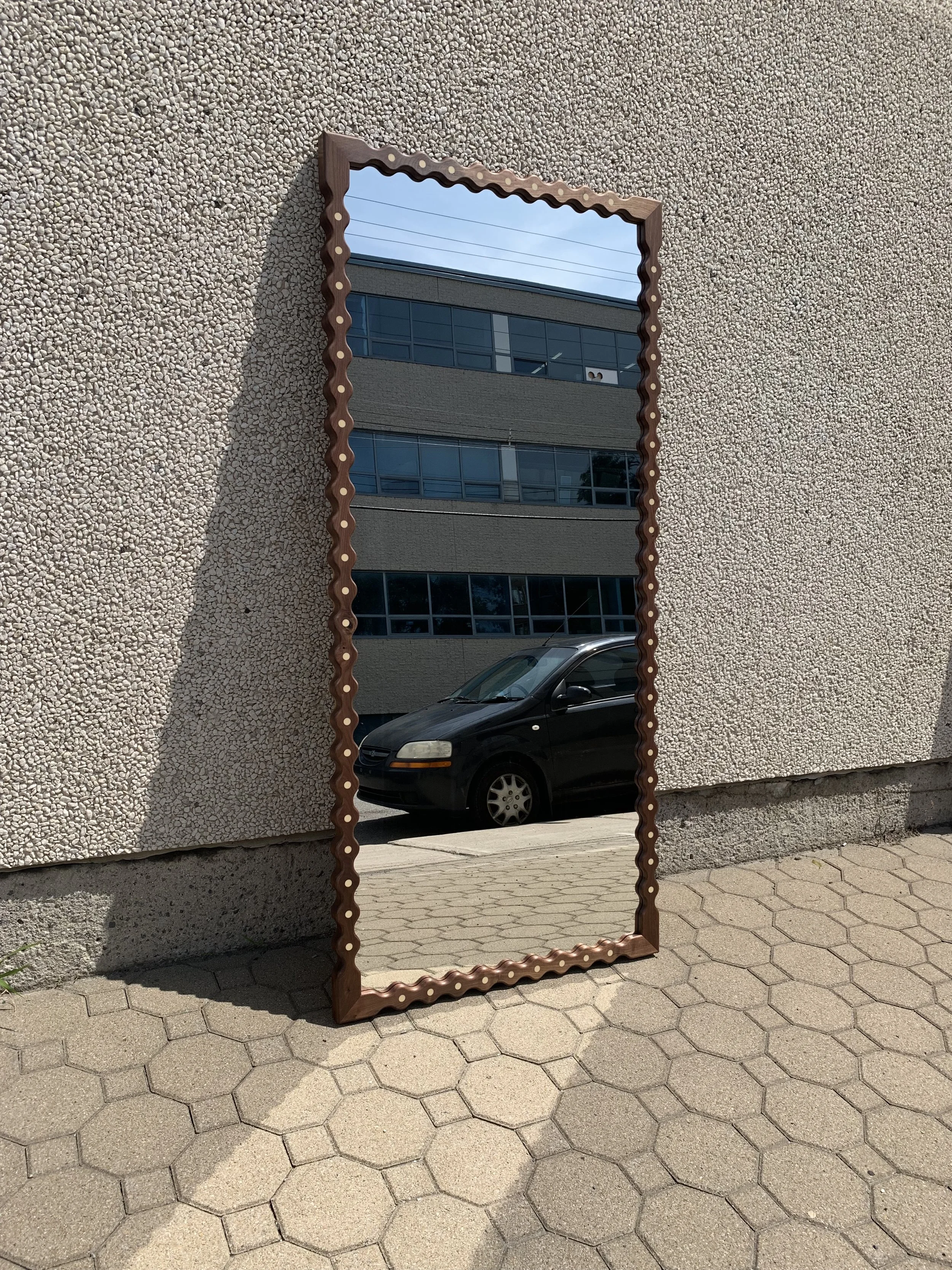 A rectangular mirror with a wavy, decorative frame leans against a textured beige wall outdoors. The mirror reflects a building with large windows and a parked black car on the street.