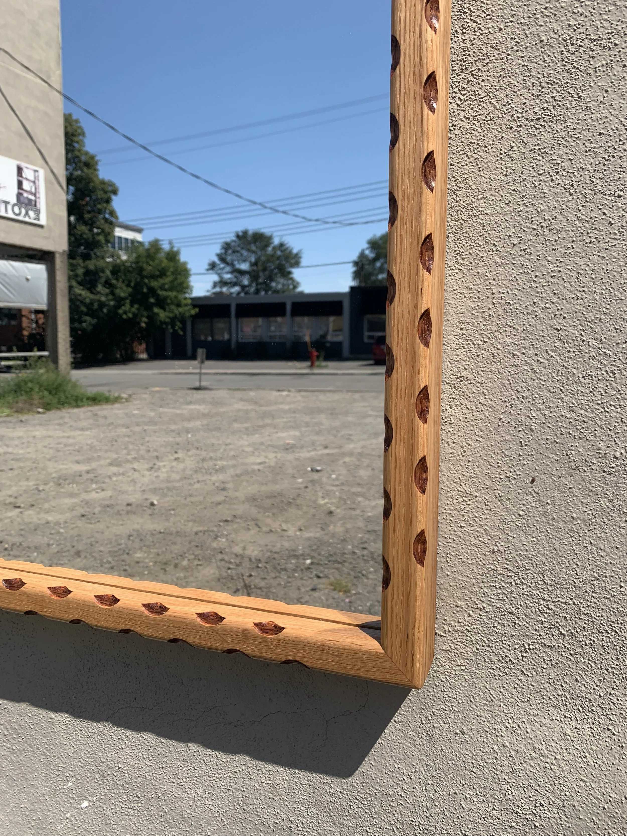 Close-up of a wooden mirror frame with carved details, reflecting an outdoor scene with a parking lot, buildings, trees, and power lines under a blue sky.