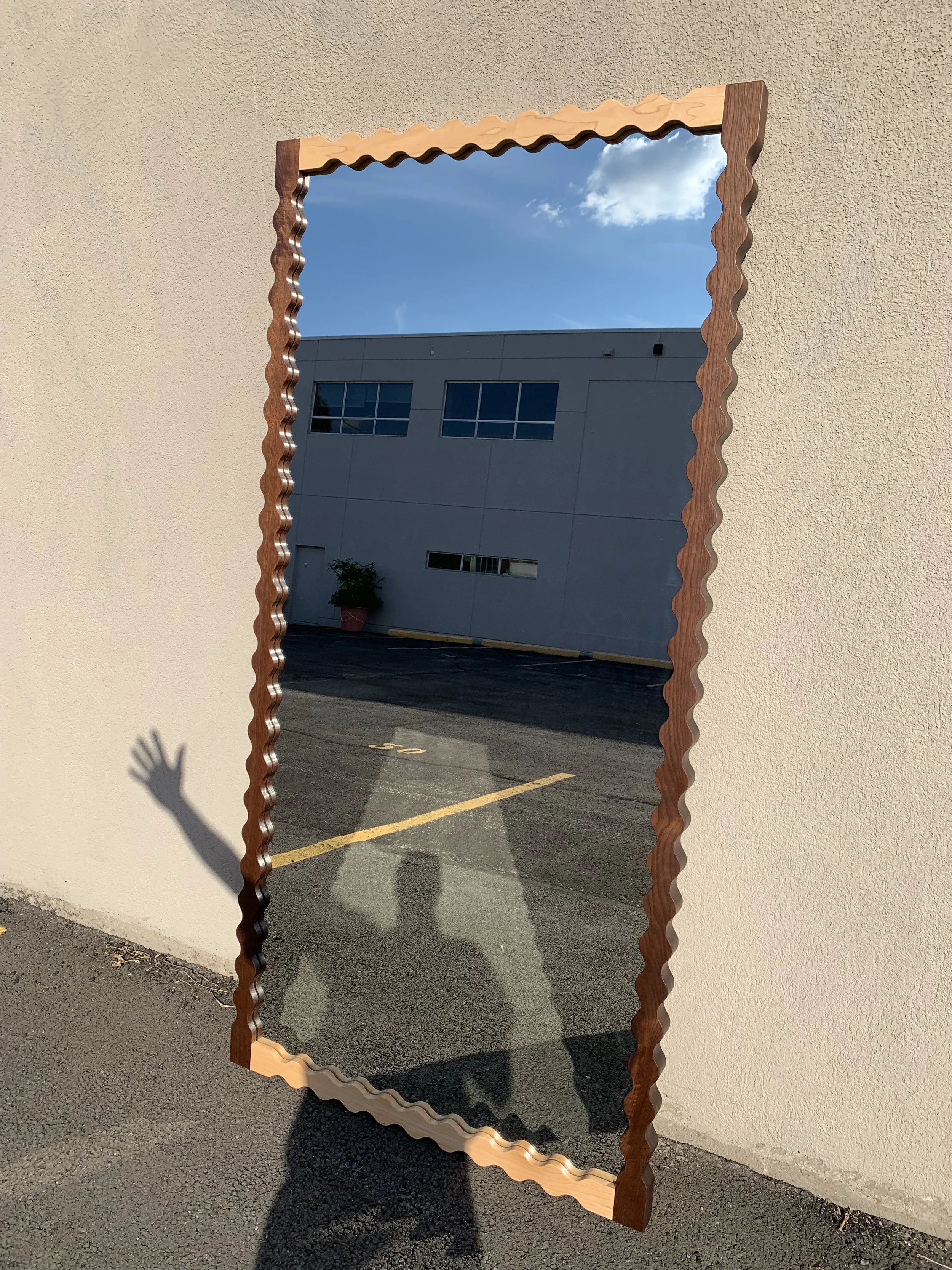 A tall mirror with a wavy wooden frame, leaning against a textured beige wall outdoors. The mirror reflects a blue sky with clouds, a building with windows, and a person's shadow with one arm raised.