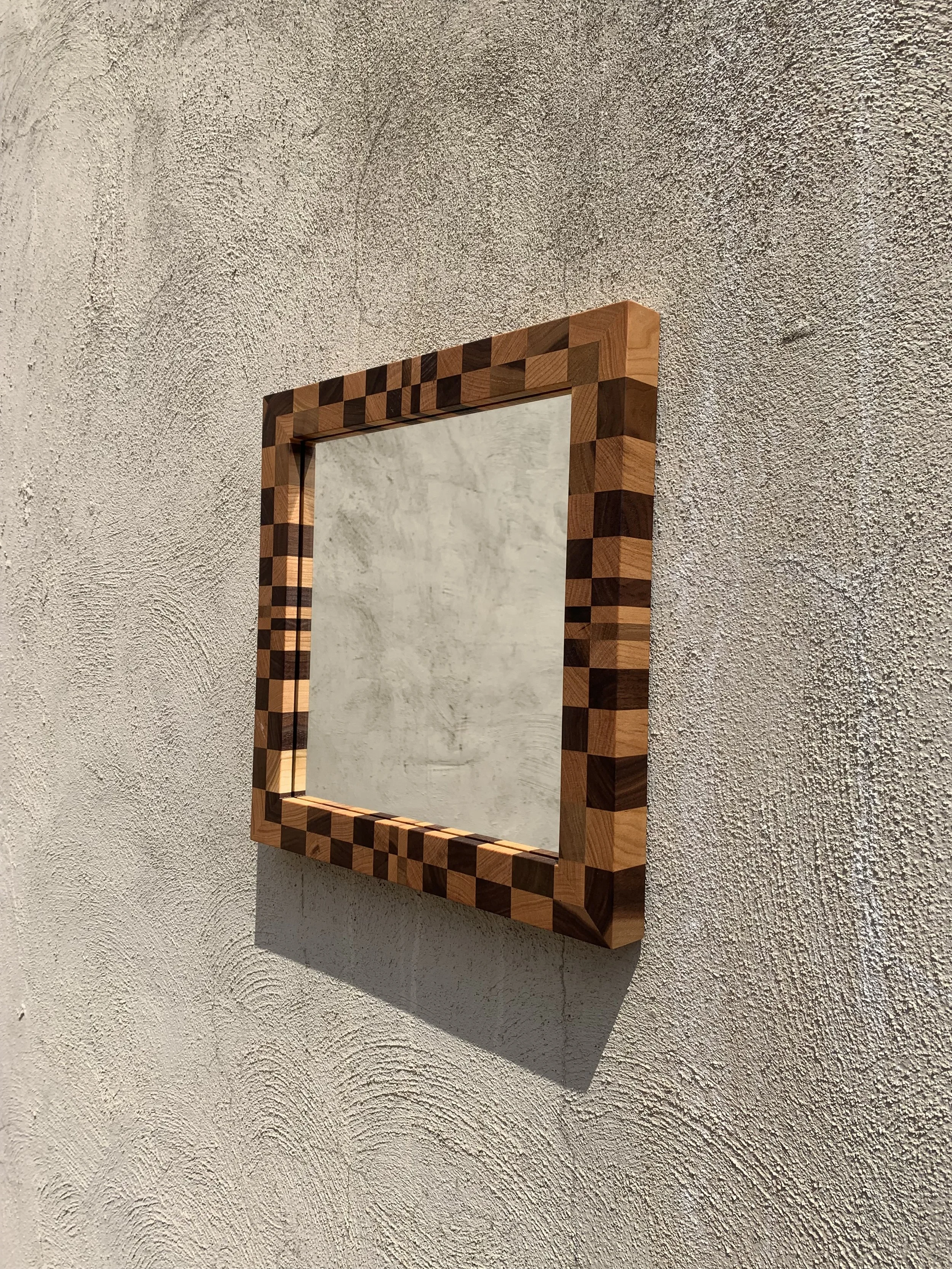 A square mirror with a wooden frame featuring a checkered pattern, mounted on a textured off-white wall.