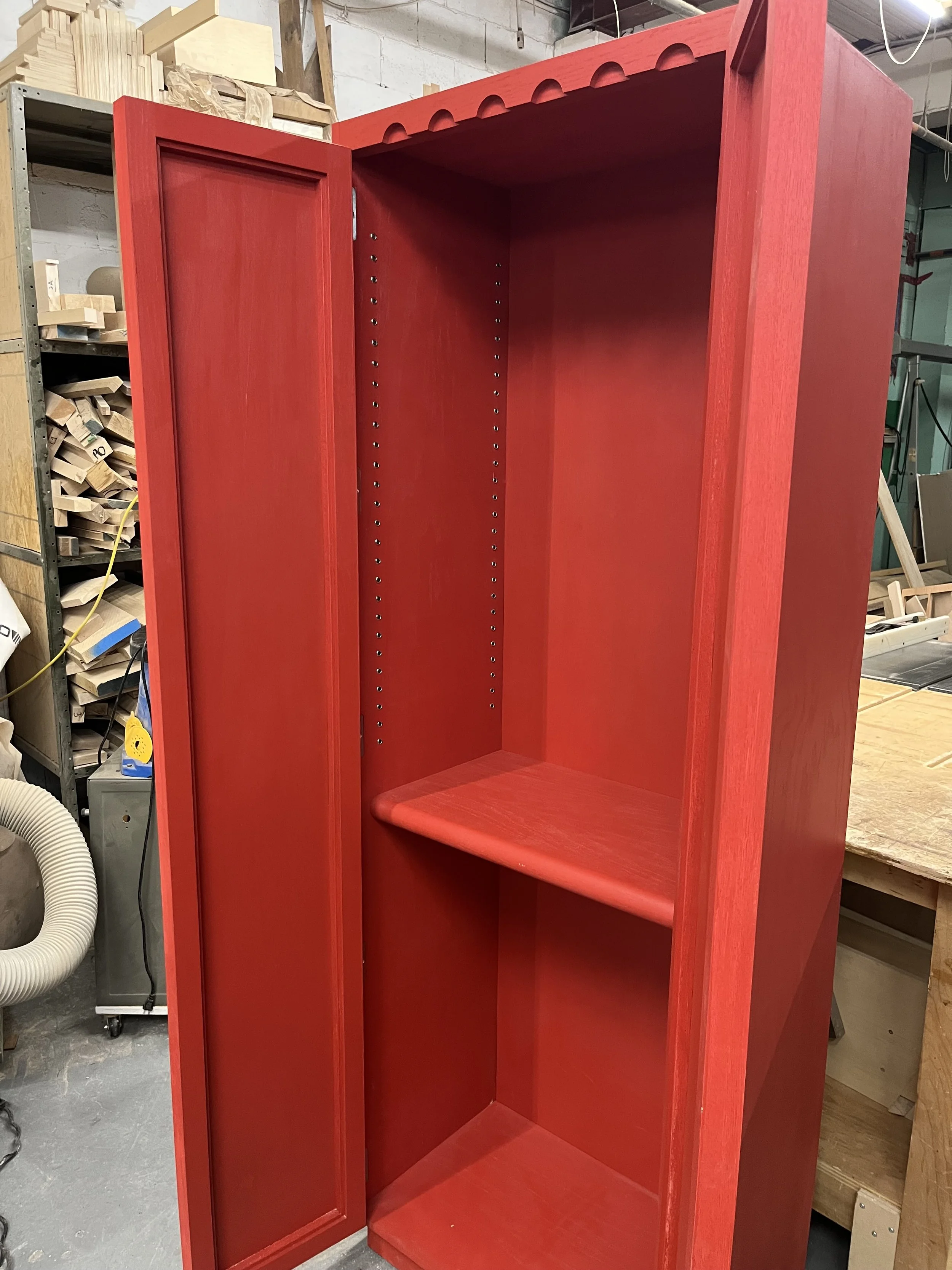 Red wooden locker with an open door inside a woodworking shop.