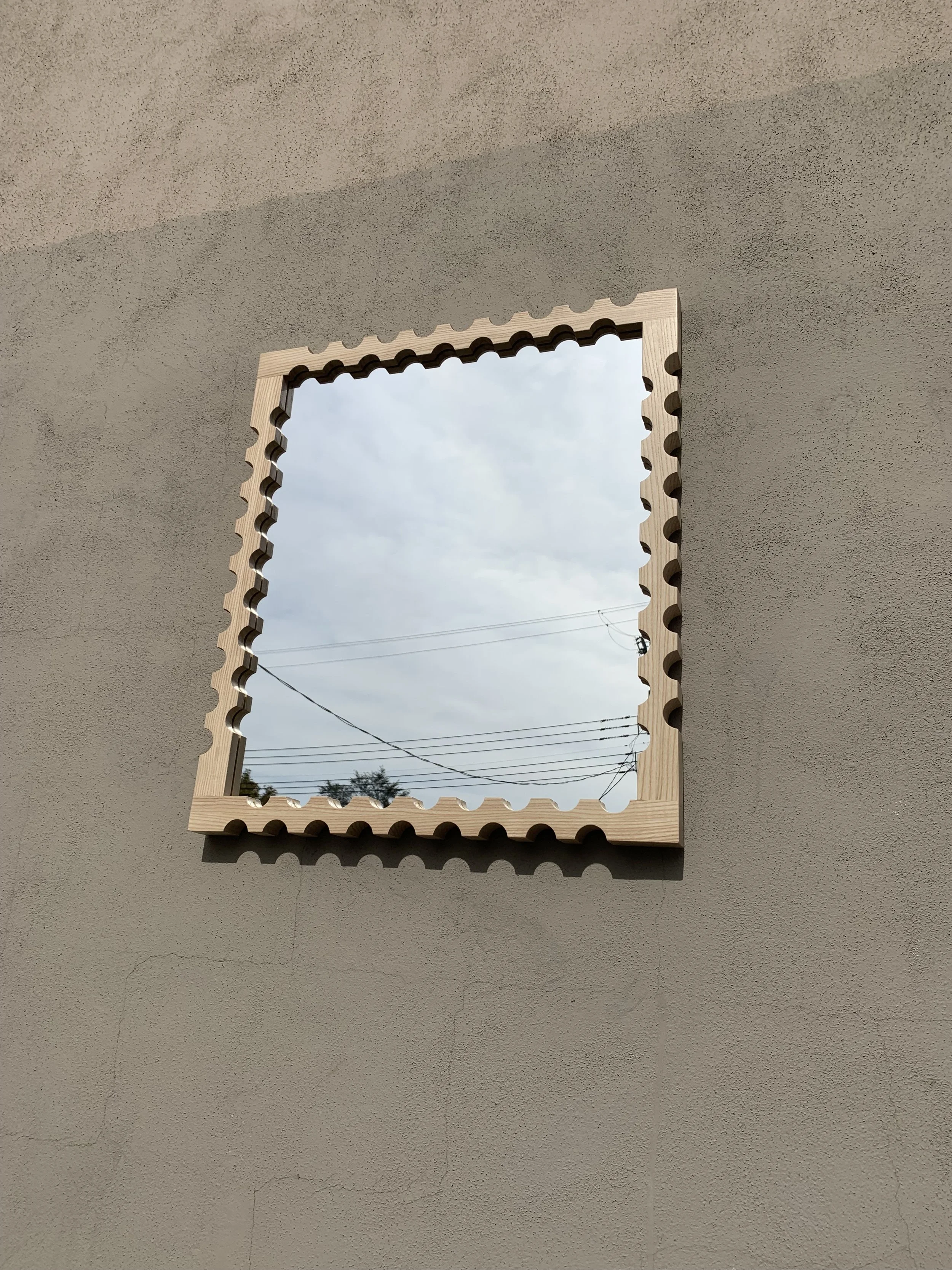 A rectangular window with a wooden frame that has a scalloped edge design, set into a beige wall with a rough texture. Outside the window, power lines and a cloudy sky are visible.