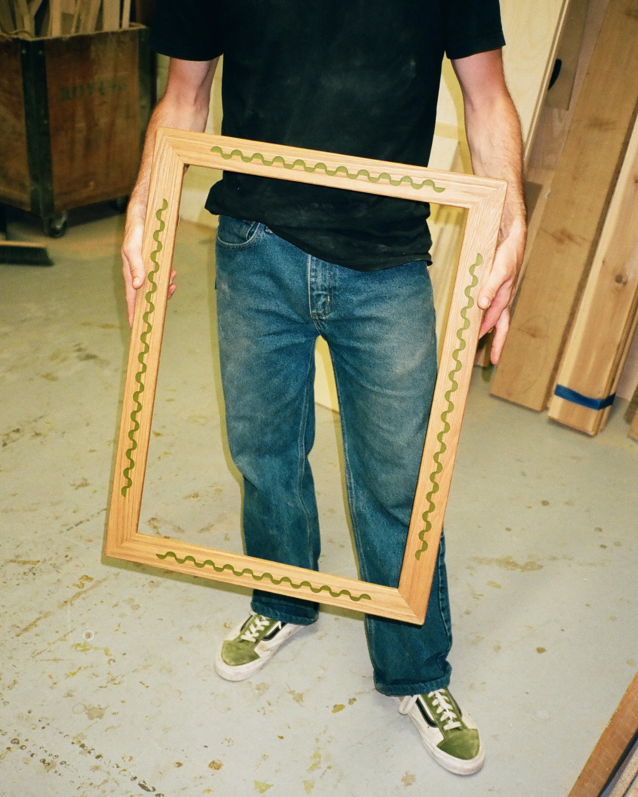 Person holding an empty wooden picture frame in a workshop with wooden planks and tools in the background.