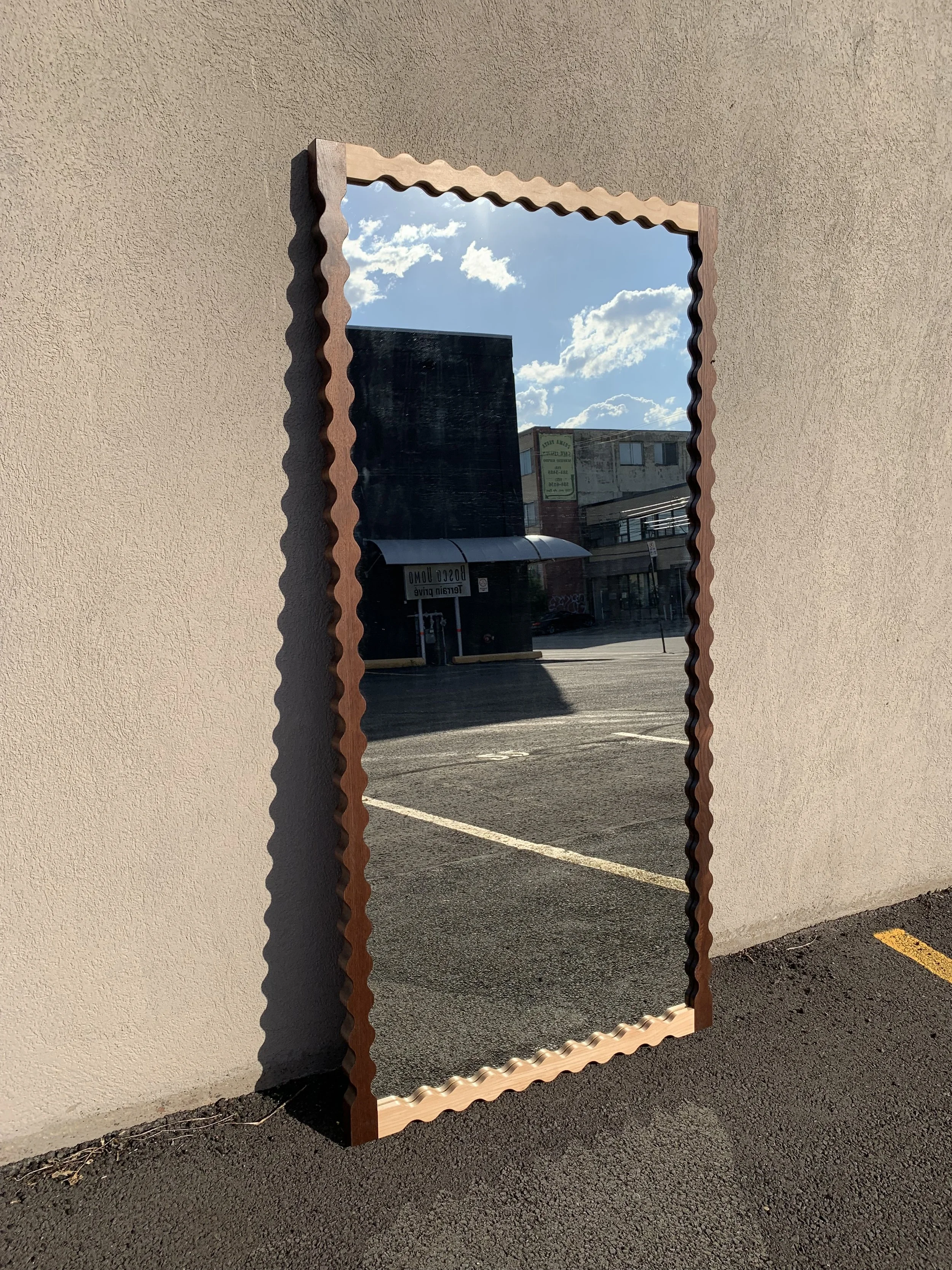 Full-length mirror with a scalloped wooden frame leaning against a beige wall outside, reflecting a parking lot, a black building, and a partly cloudy sky.