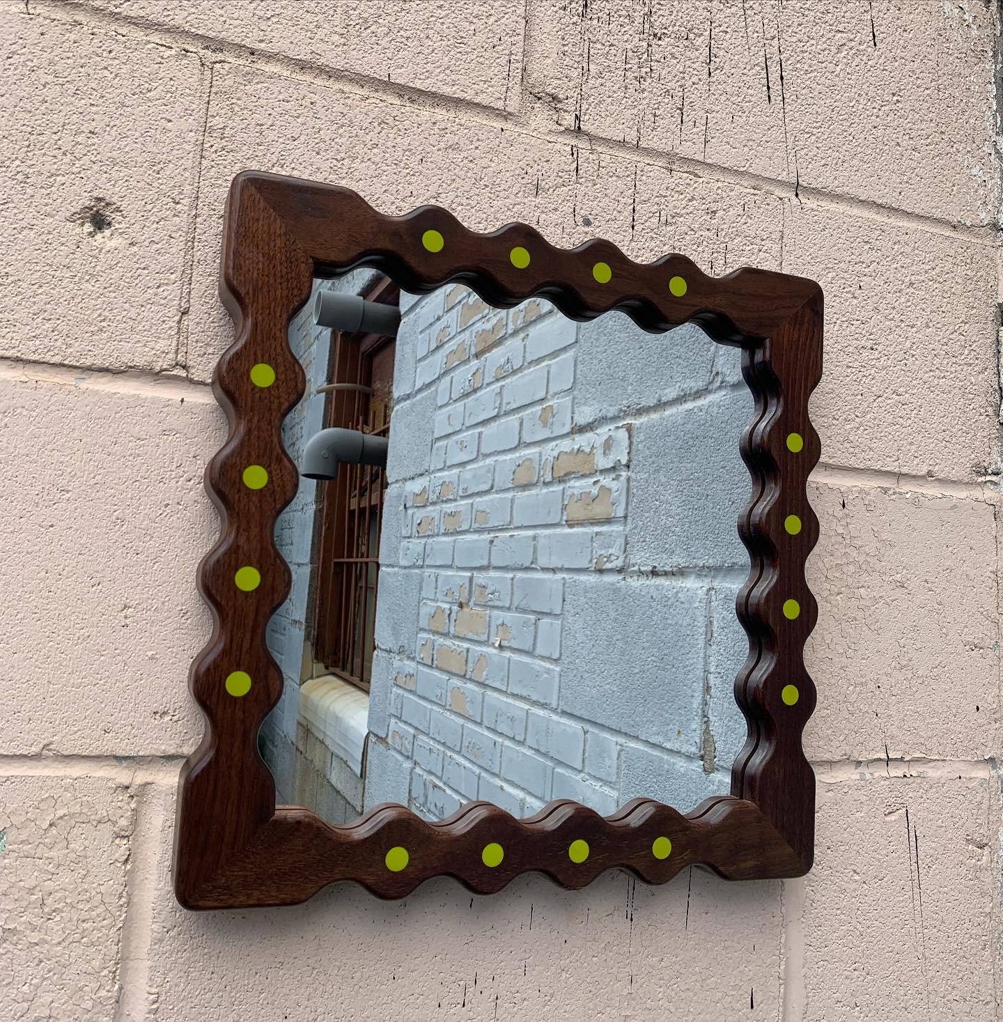 A square wooden mirror frame with wavy edges and green dots on each side, mounted on a pink concrete block wall with bricks and a window reflected in the mirror.