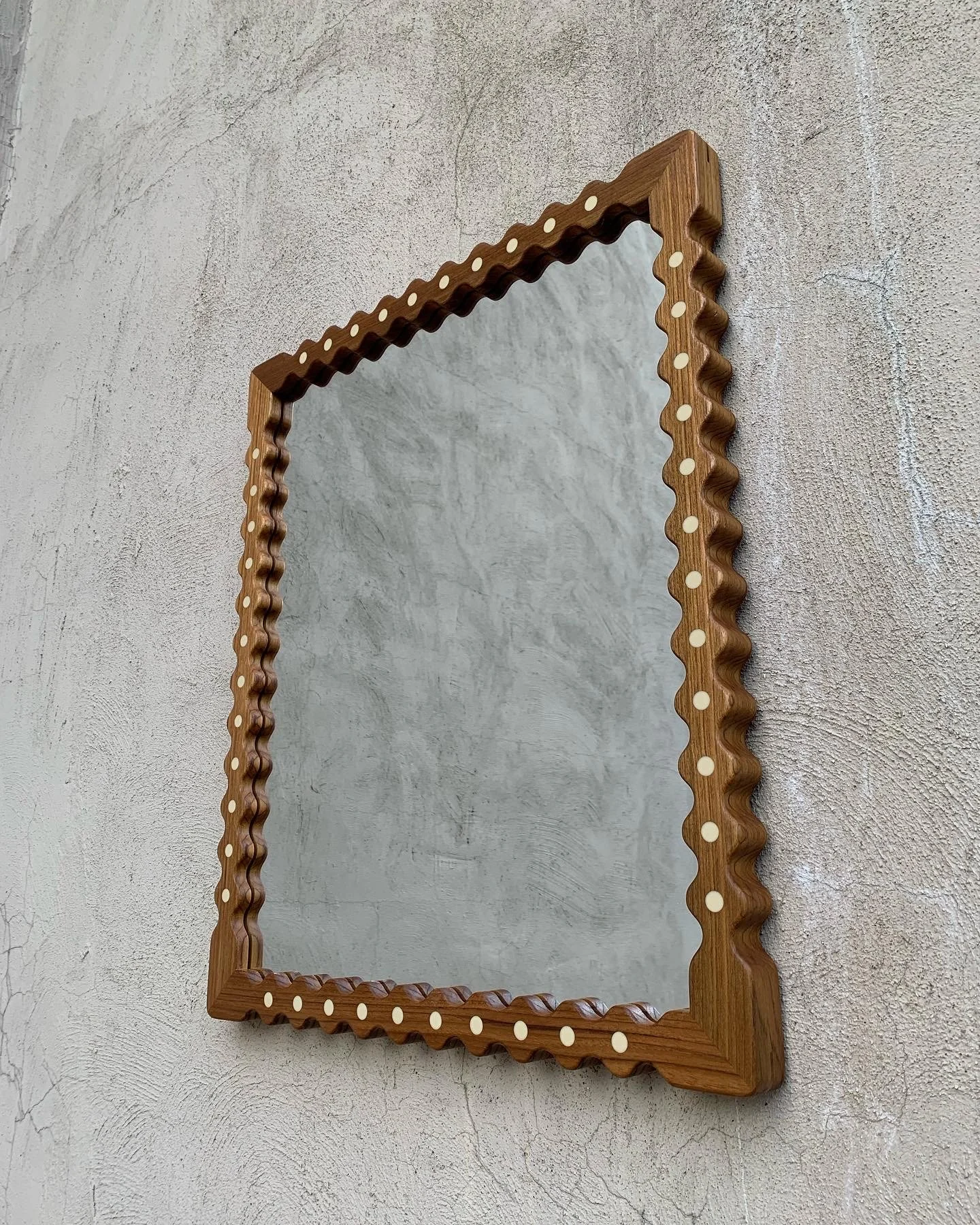 A rectangular mirror with a wooden frame featuring a scalloped design and white dotted accents, mounted on a textured wall.