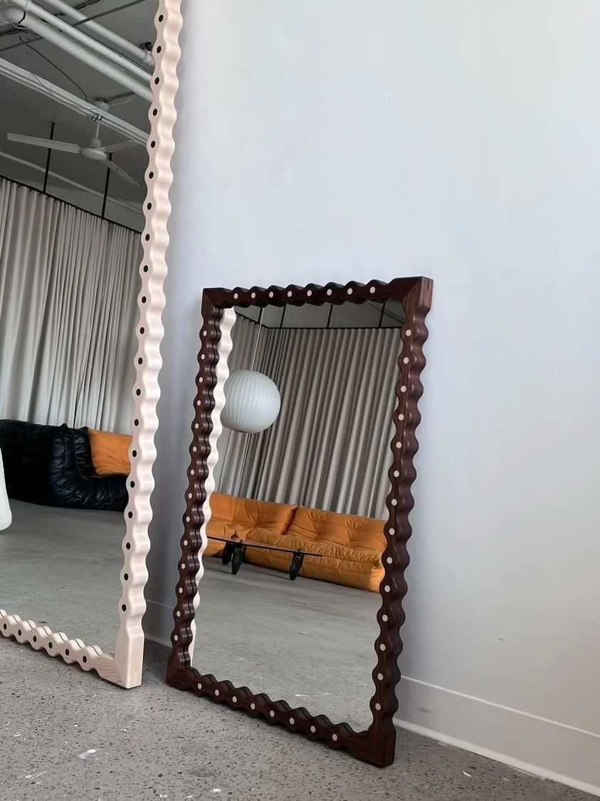 Two large decorative mirrors with scalloped edges, one white and one dark wood, leaning against a plain white wall. The mirrors reflect a curtain, a round paper lantern, and a tan sofa.