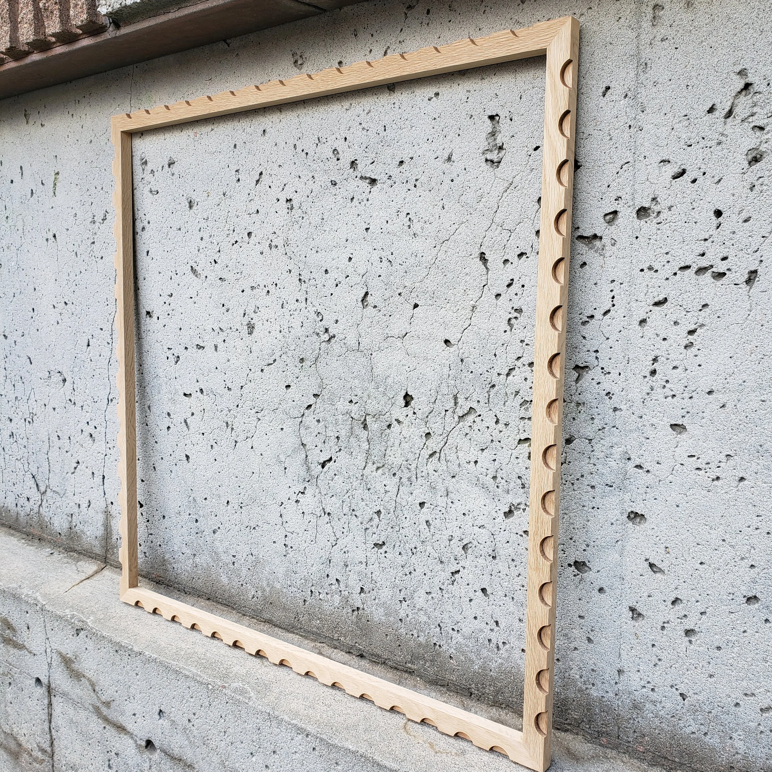 A wooden frame with evenly spaced circular cutouts leaning against a concrete wall with cracks and rough texture.