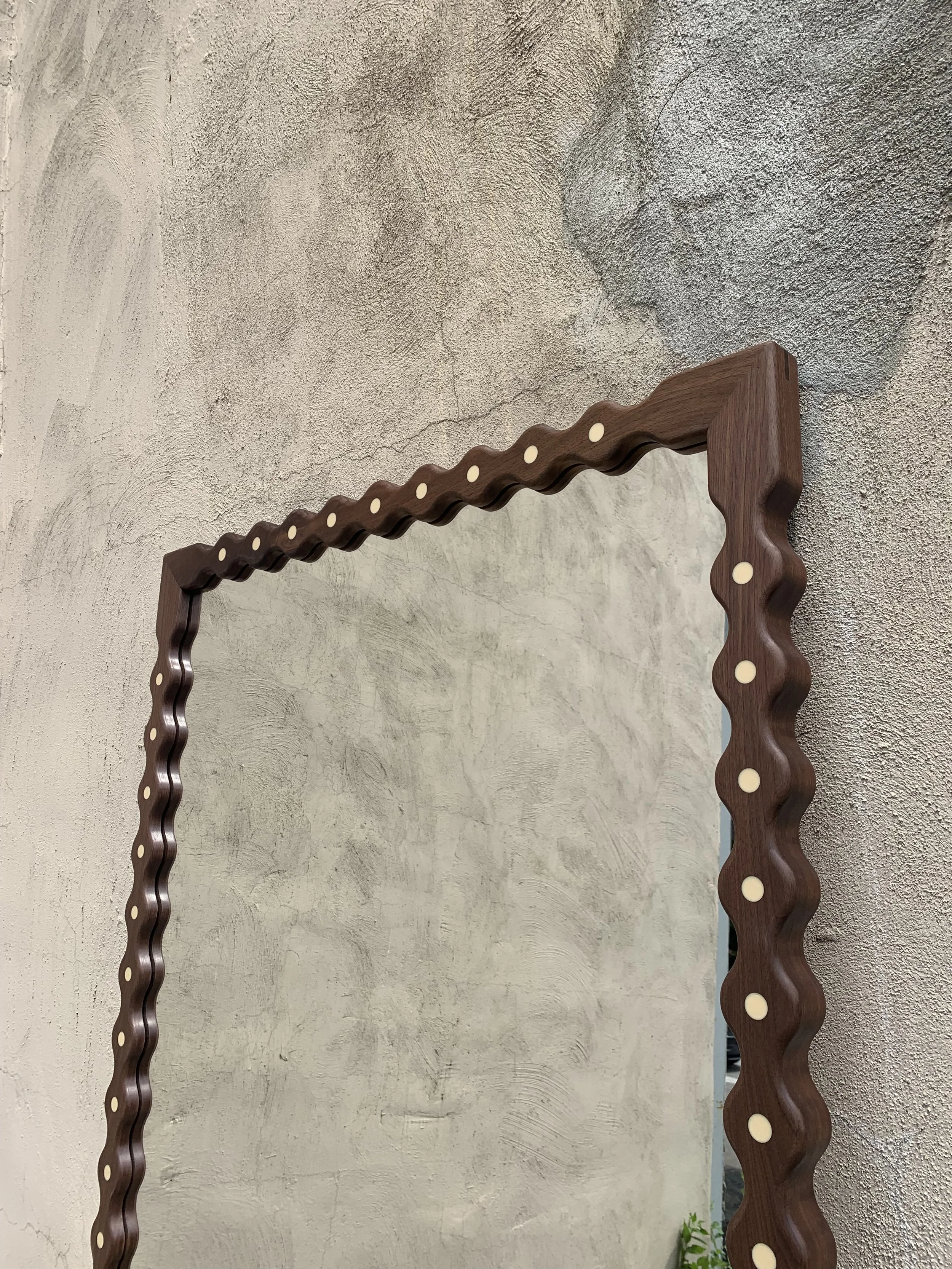 Decorative wooden mirror with a wavy, scalloped frame decorated with circular inlays, mounted on a textured wall.