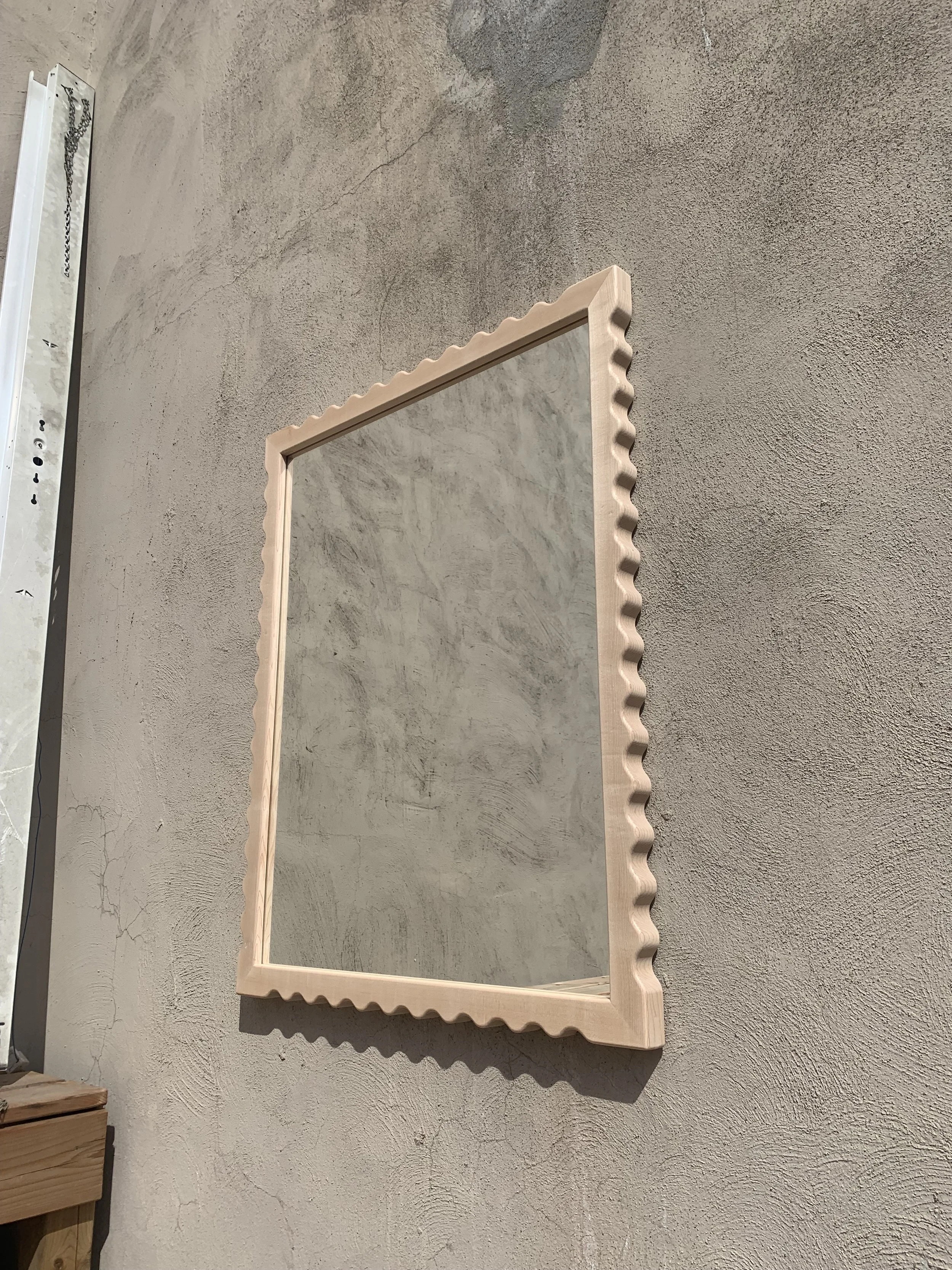 A rectangular mirror with a wavy, textured beige frame hanging on a rough, gray concrete wall.