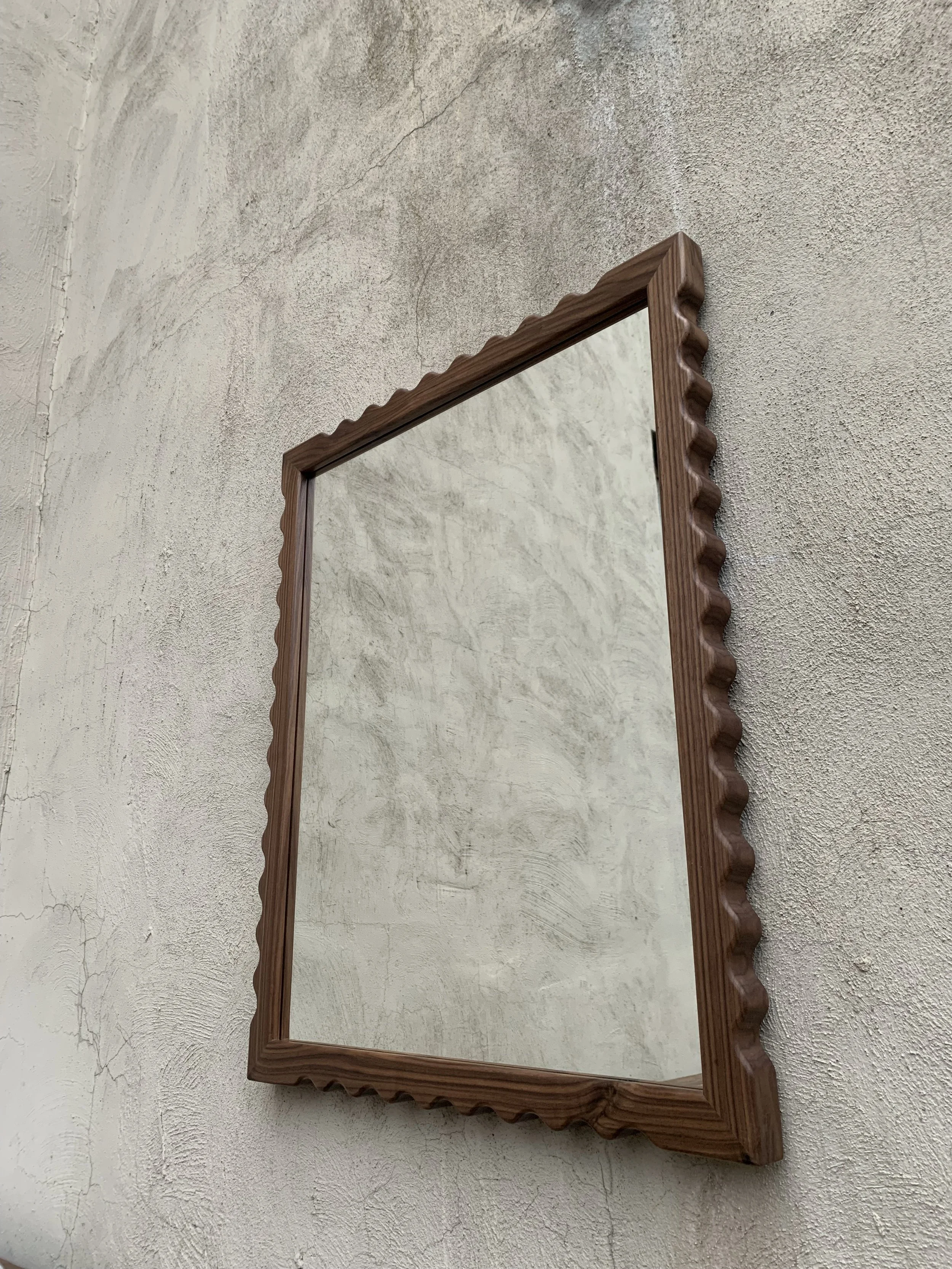 A rectangular mirror with a wavy wooden frame hanging on a textured white wall.