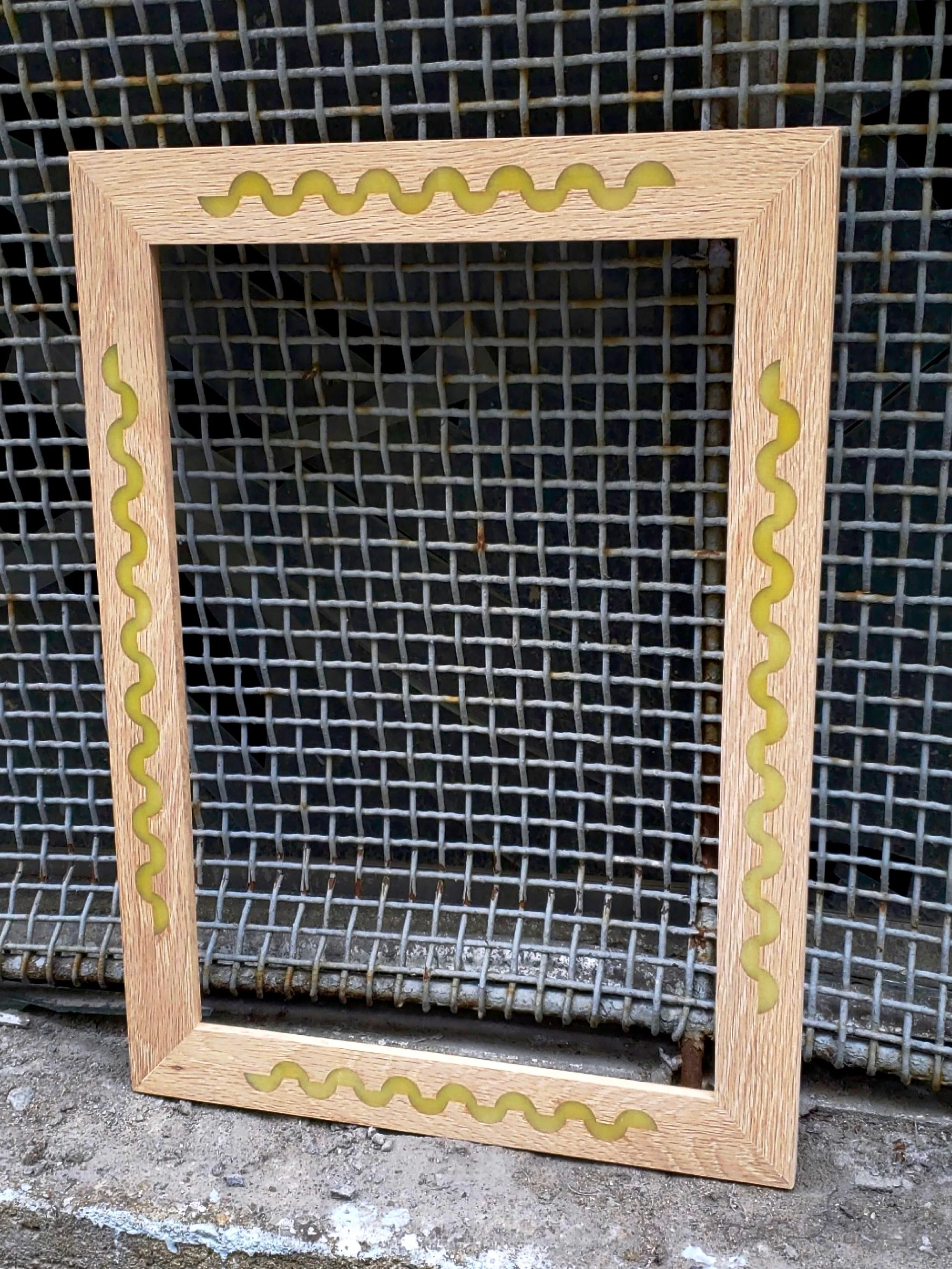 Wooden picture frame with yellow wavy design on each side, placed against a metal wire mesh background.
