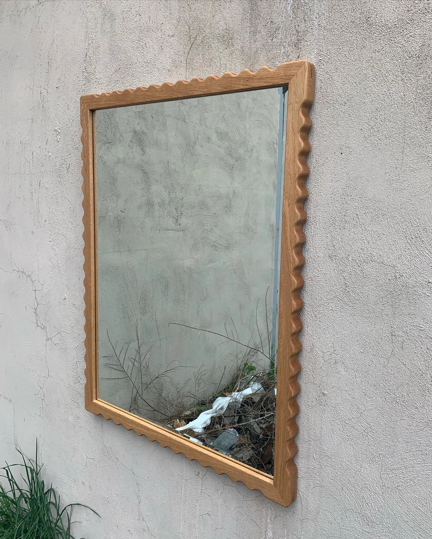 A rectangular mirror with a decorative wooden frame hanging on a textured beige wall. The reflection shows a concrete wall with some cracks and debris, including trash and branches, near the bottom of the mirror.