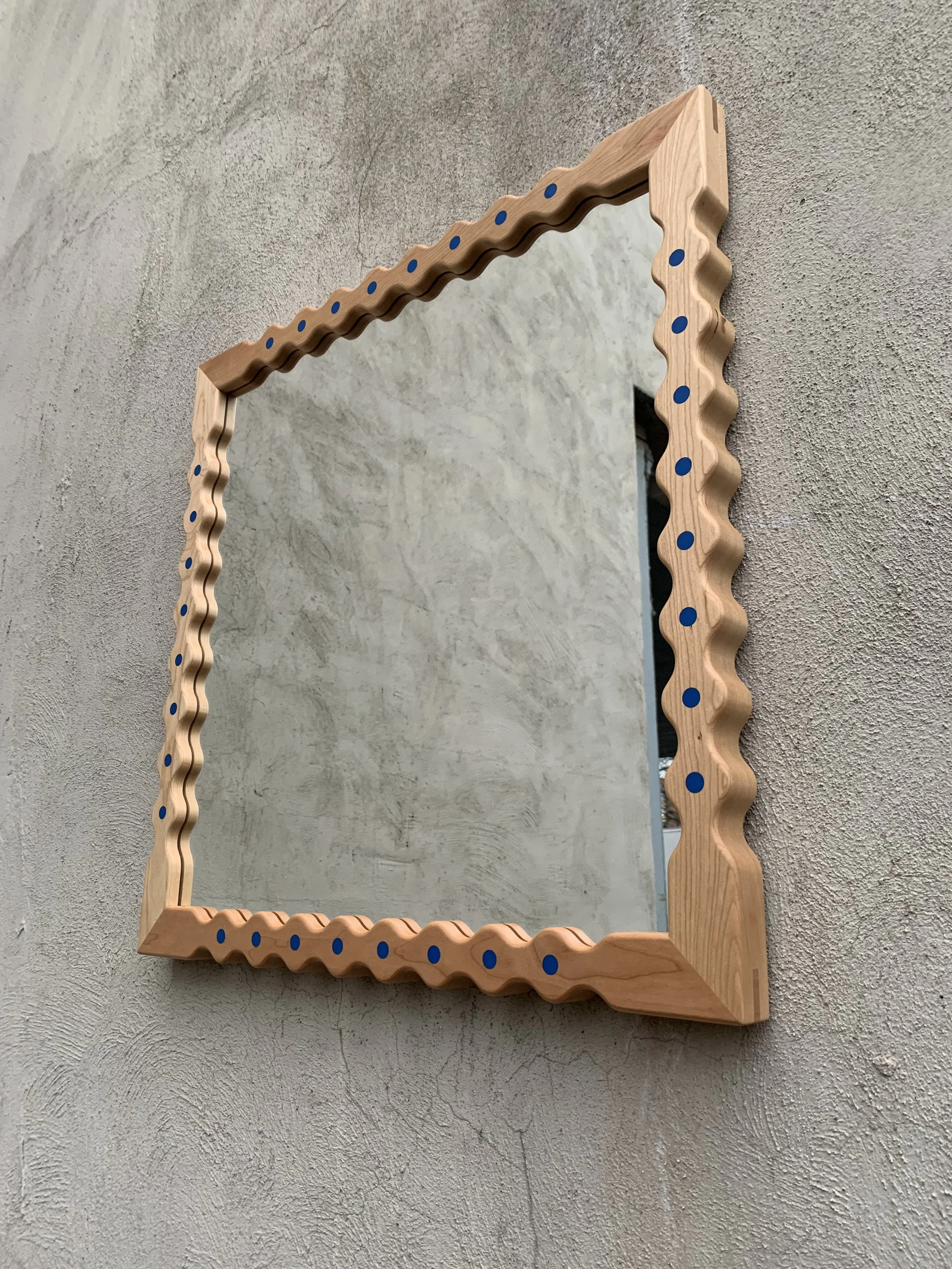 Wooden-framed mirror with wavy edges and blue dots on the frame, hung on a textured gray wall.