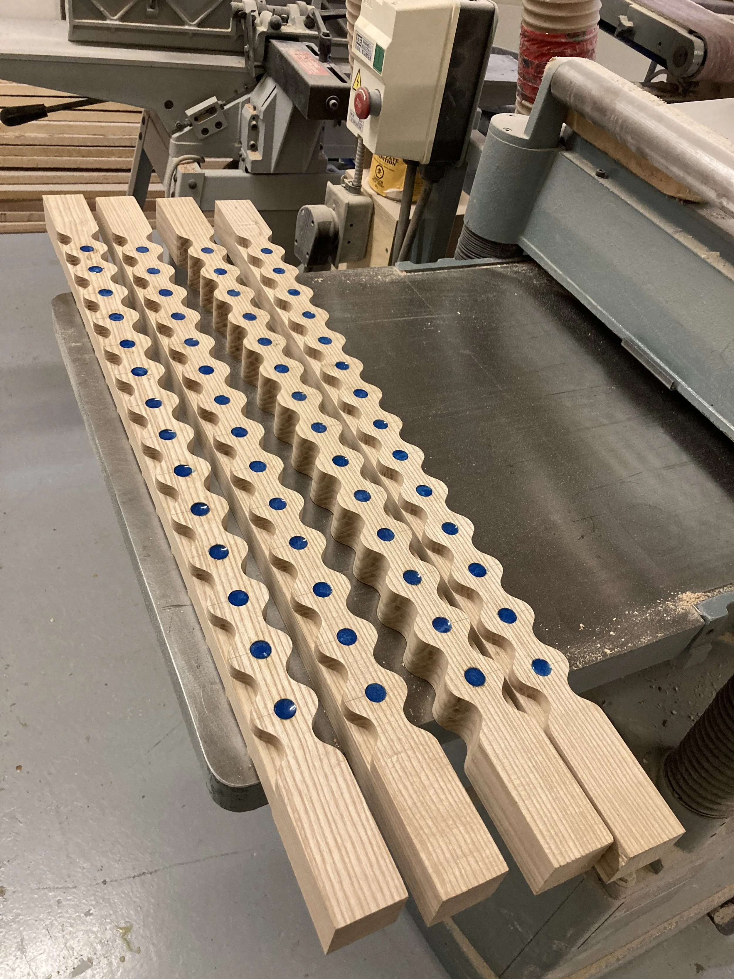 Four wooden pieces with wavy cutouts and blue circular inserts, placed on a woodworking table in a workshop.