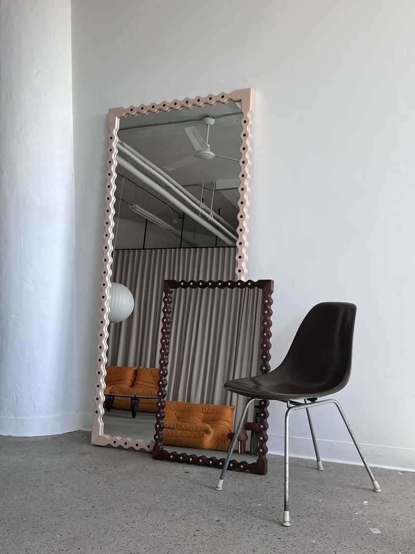 A black chair and two large decorative mirrors leaning against a white wall. One mirror has a pink frame, the other has a dark brown frame. Their reflections show a ceiling fan, a white light fixture, and orange cushions on a sofa.