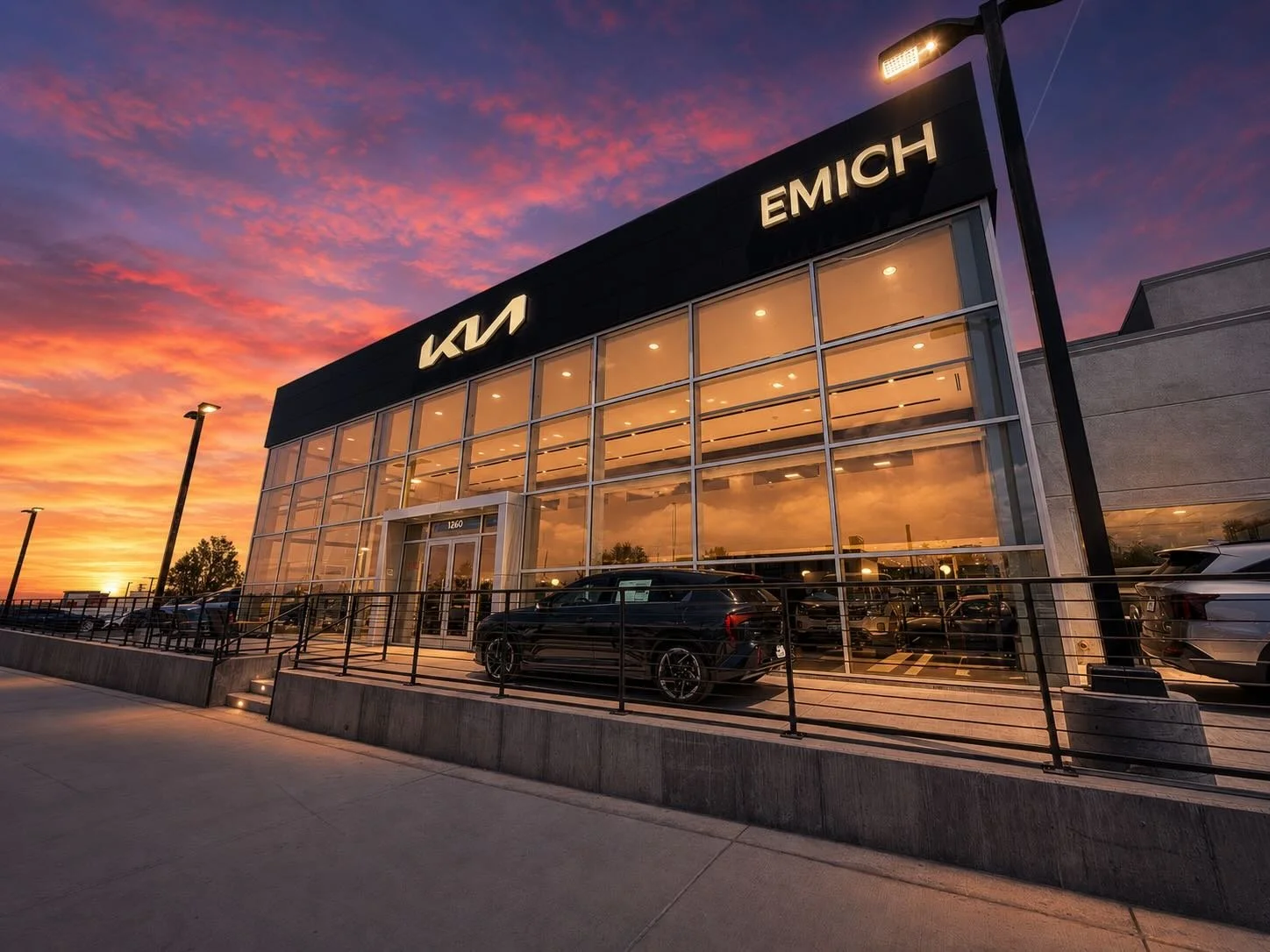 Modern Kia car dealership building during sunset with illuminated signage, large glass windows, and parked cars outside.
