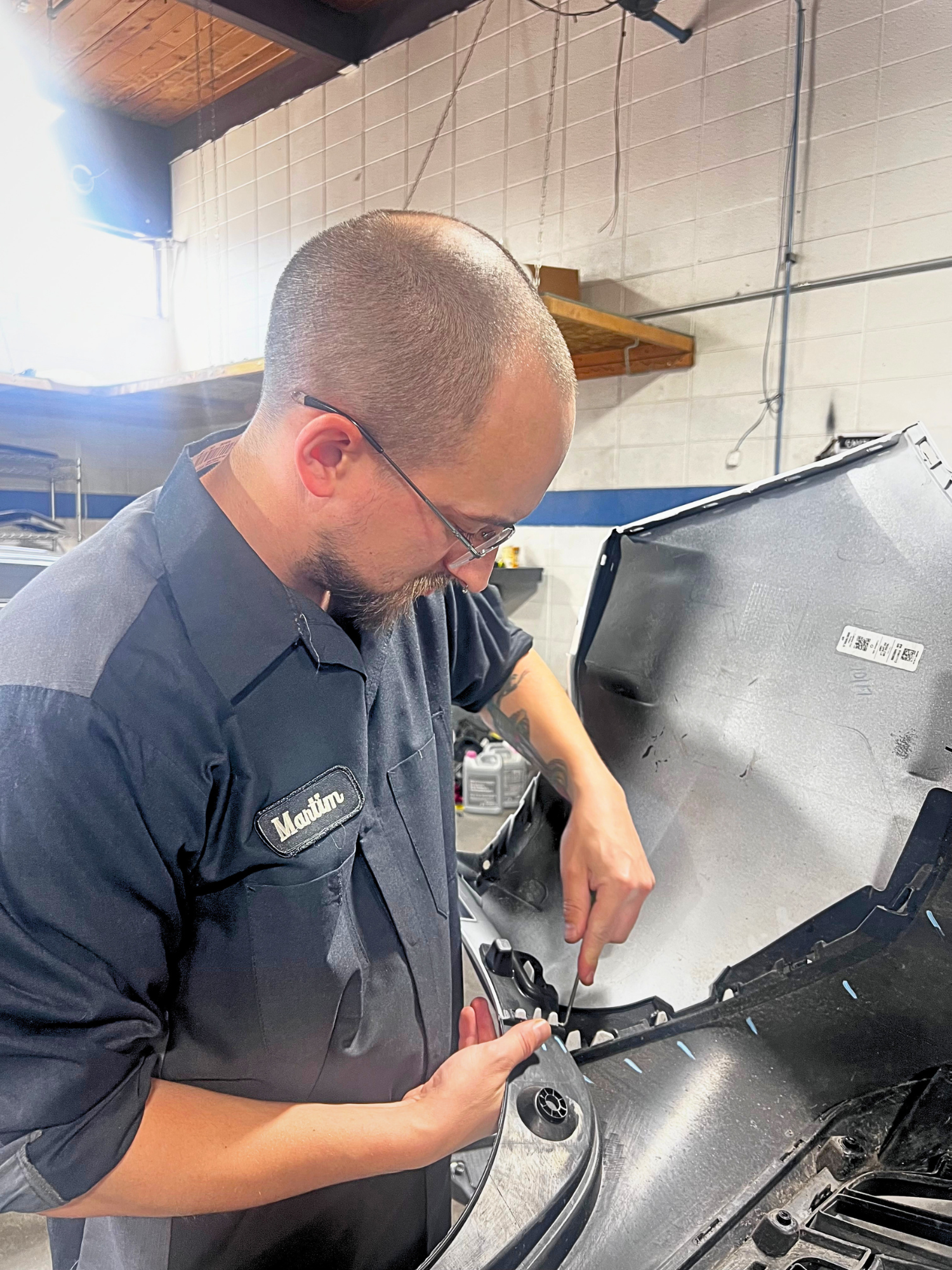 A mechanic lying on a creeper under a silver car in a garage, inspecting or repairing the vehicle.