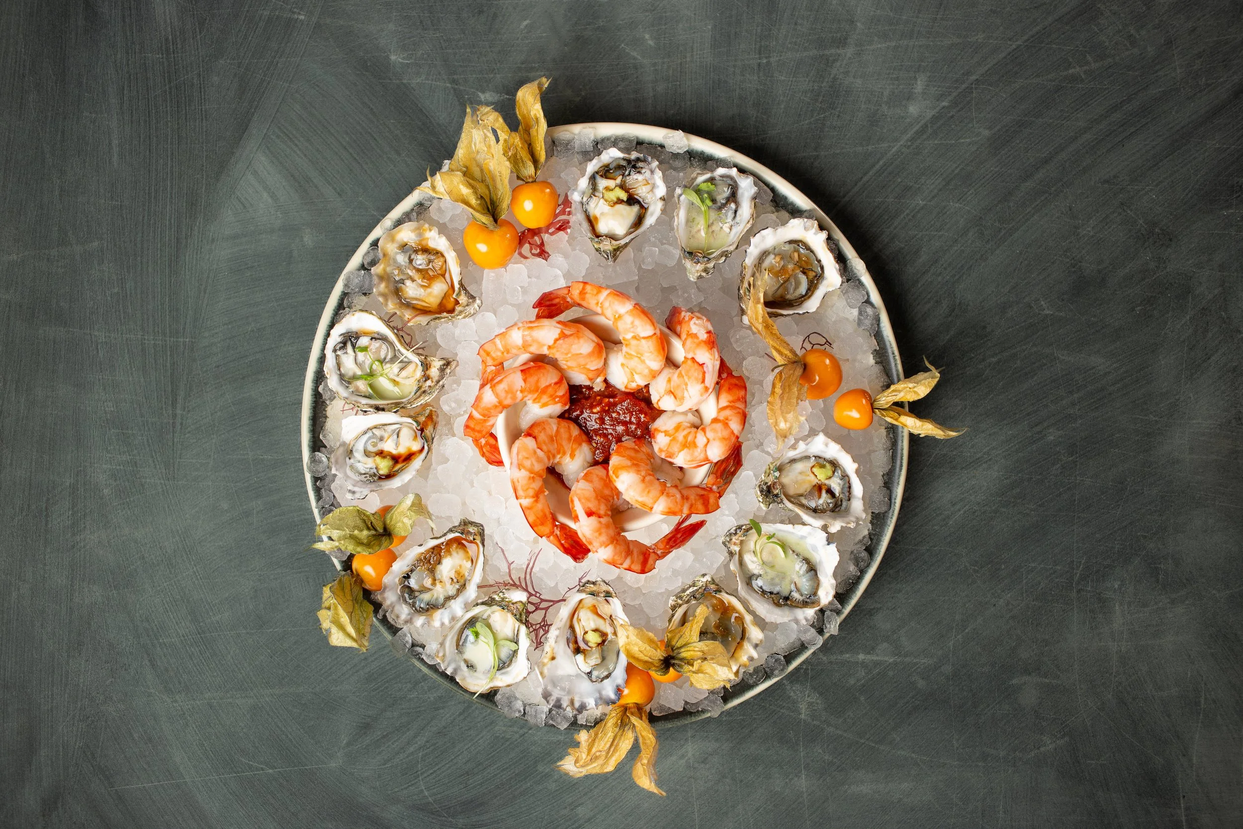 A round platter of oysters on ice, garnished with small orange cherry tomatoes and dried leaves, with the oysters arranged around a central cluster of cooked shrimp on a bed of crushed ice against a dark background.